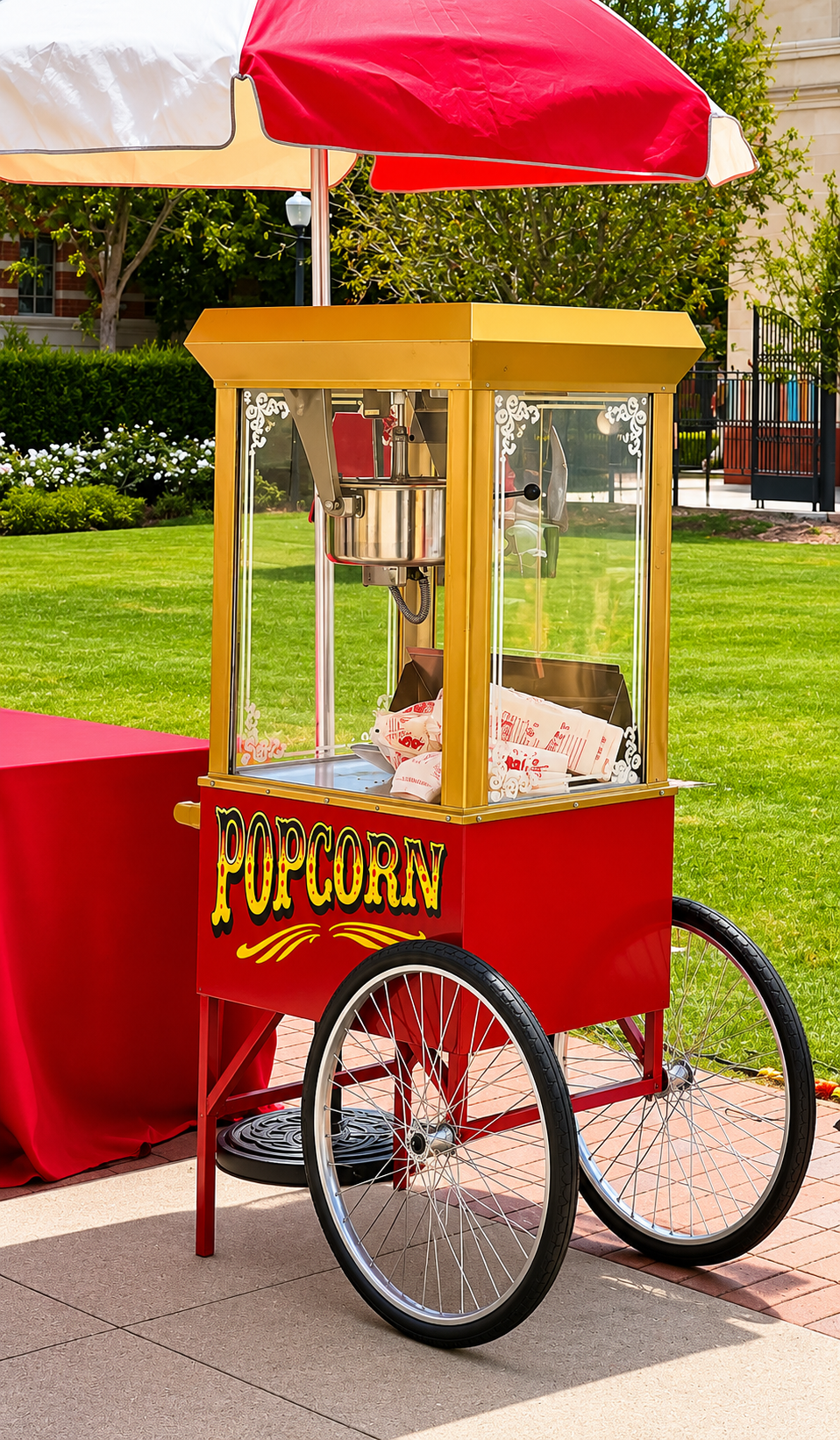 A red and yellow popcorn cart with two large black wheels, a red umbrella, and a glass enclosure, situated on a sidewalk with a grassy park and trees in the background.