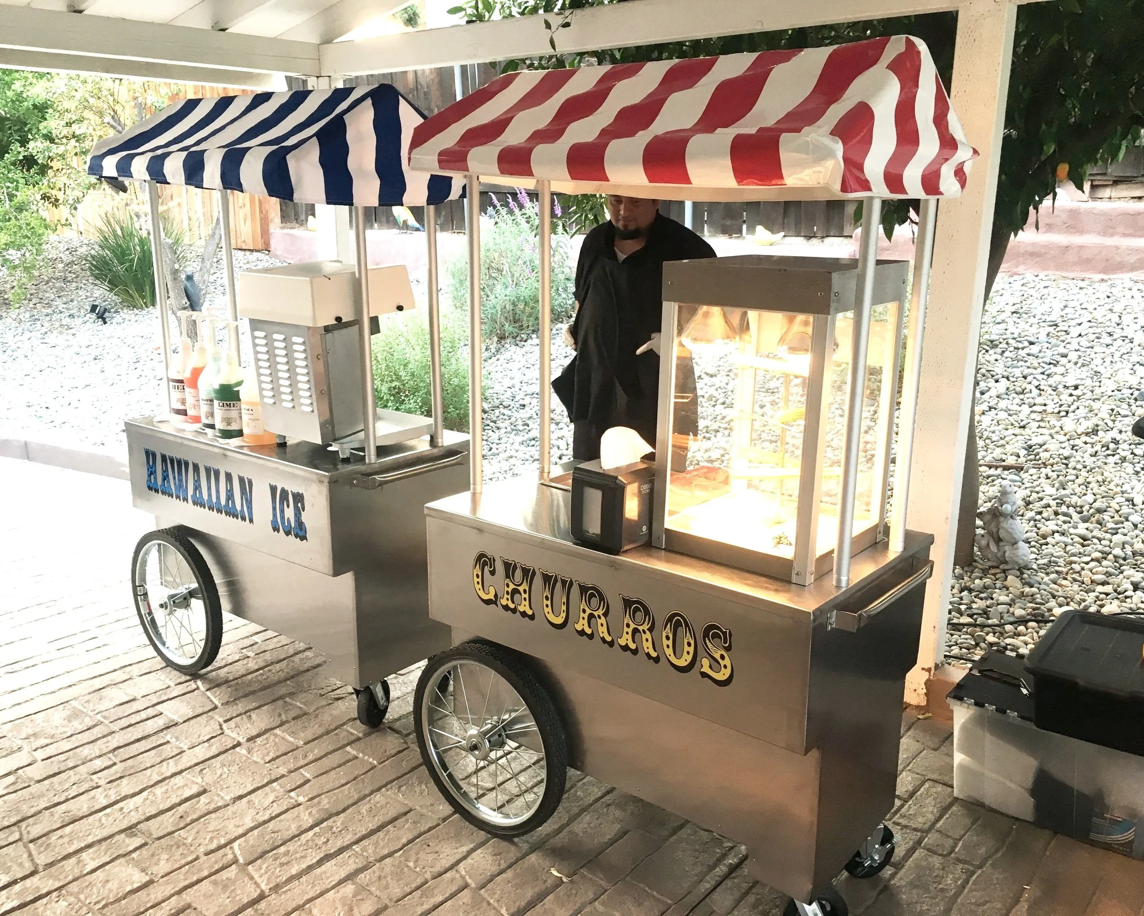 A mobile popcorn cart with striped red and white and blue and white umbrellas, situated on a brick pathway with greenery in the background. The cart has a sign that reads 'Hawaiian Ice' and another sign that says 'Popcorn.' A man is standing behind the cart.