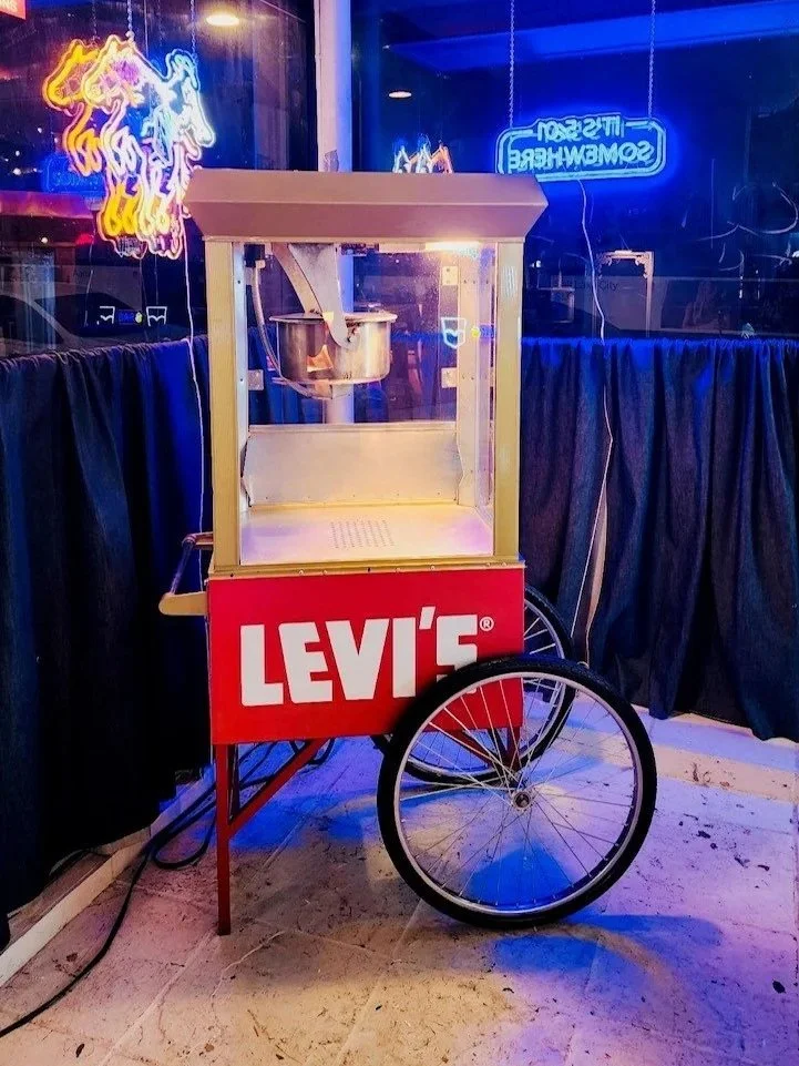 A popcorn cart with a red Levi's logo on the front, located inside a venue with neon signs and black curtains in the background.
