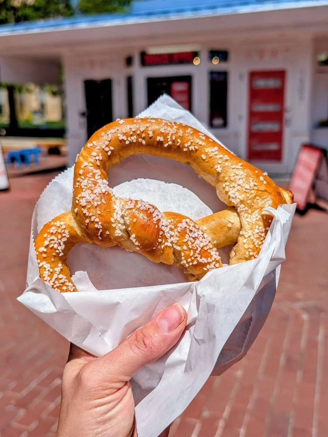 Two sugar-covered pretzels in a paper bag held in front of a food stand with a red and white facade.