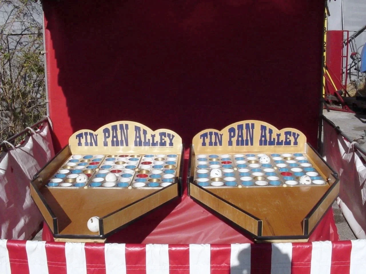 Two wooden tic-tac-toe game boards labeled 'Tin Pan Alley' on red and white striped table at outdoor fair.