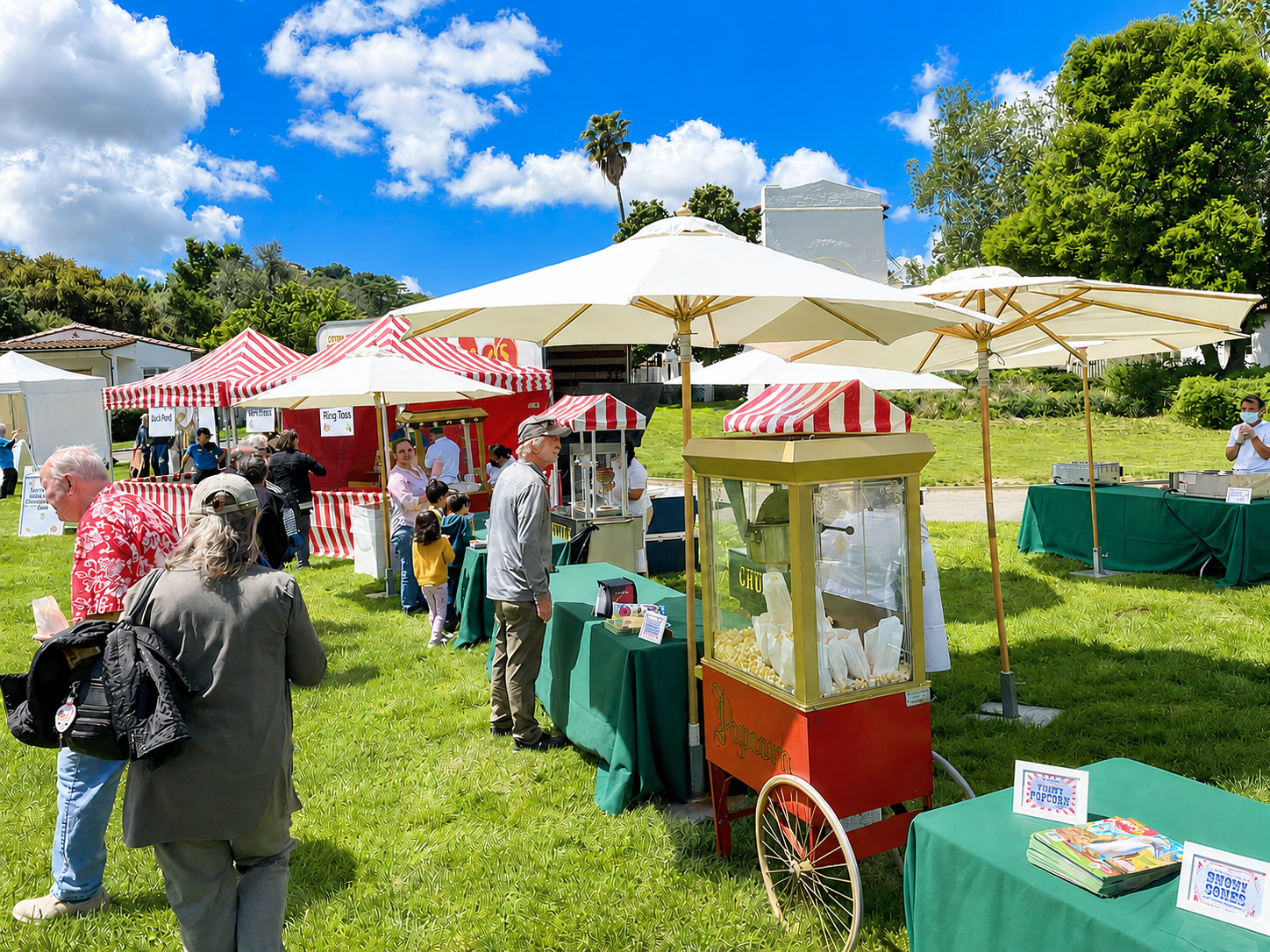 Outdoor food festival with food vendors under striped tents and umbrellas, people in line buying snacks, a popcorn cart, and a grassy area with trees and a blue sky with clouds