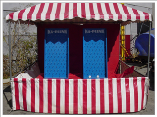 An outdoor carnival game booth with a red and white striped canopy and sides, featuring two blue paddles or game panels, set up in an open area with some trees and equipment in the background.