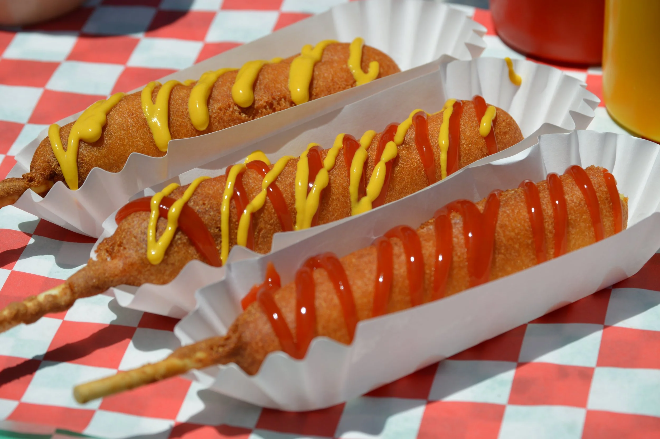 Close-up of three fried fish fillets topped with yellow mustard and red ketchup, served in white paper trays on a red-and-white checkered tablecloth.