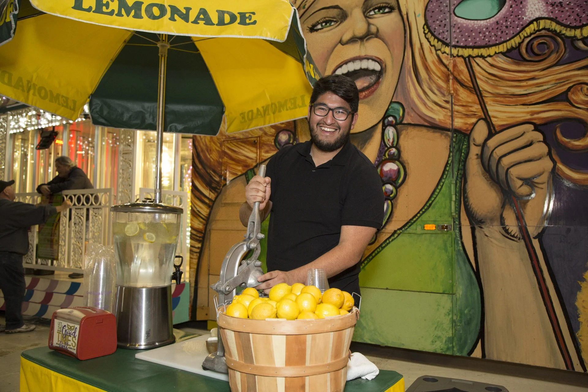 A man with glasses and a beard smiling while squeezing lemons at a lemonade stand. Behind him is a colorful mural of a woman holding an umbrella. The stand has a basket of lemons and a dispenser of lemonade with lemon slices.