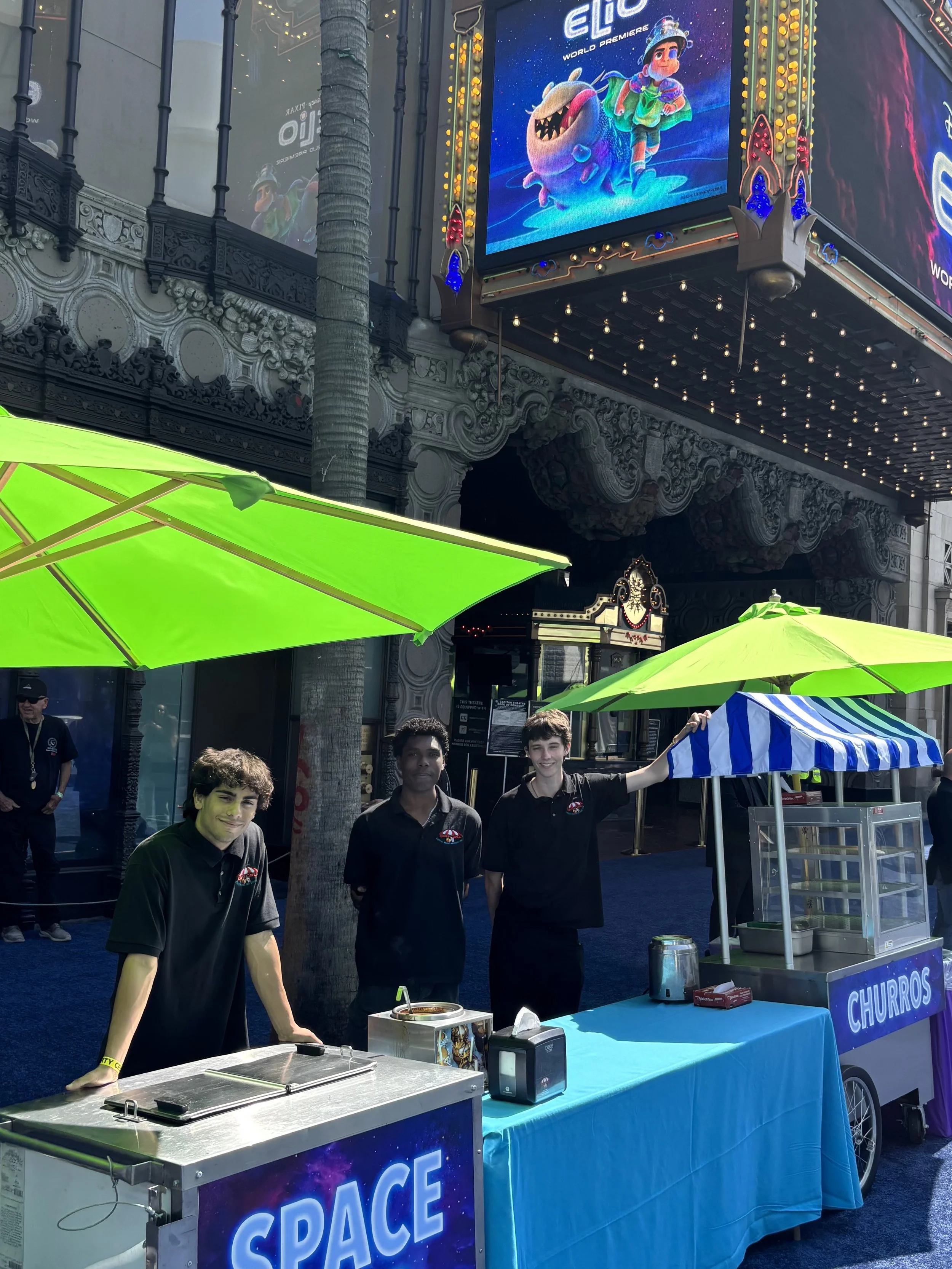 Three teenage boys working at a churro stand with a blue tablecloth and a sign that says 'Churros', outdoors on a city street with bright green umbrellas and a large ornate building in the background. Digital billboard advertising animated characters