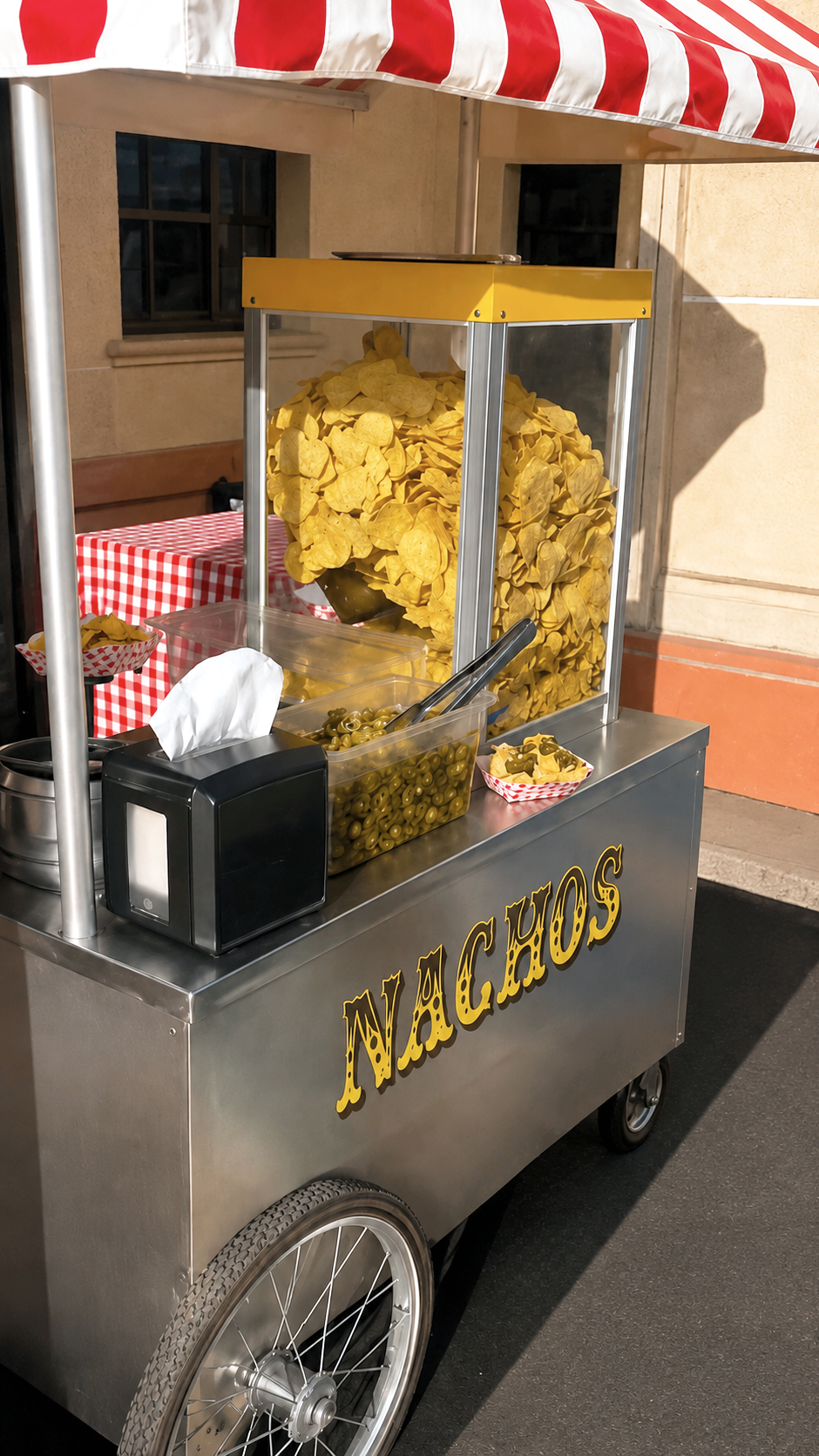 A street food cart selling potato chips and green olives, with a large bag of potato chips and a bowl of olives visible. The cart has the word 'NACHOS' written on it and is set up outdoors near a beige building with windows.