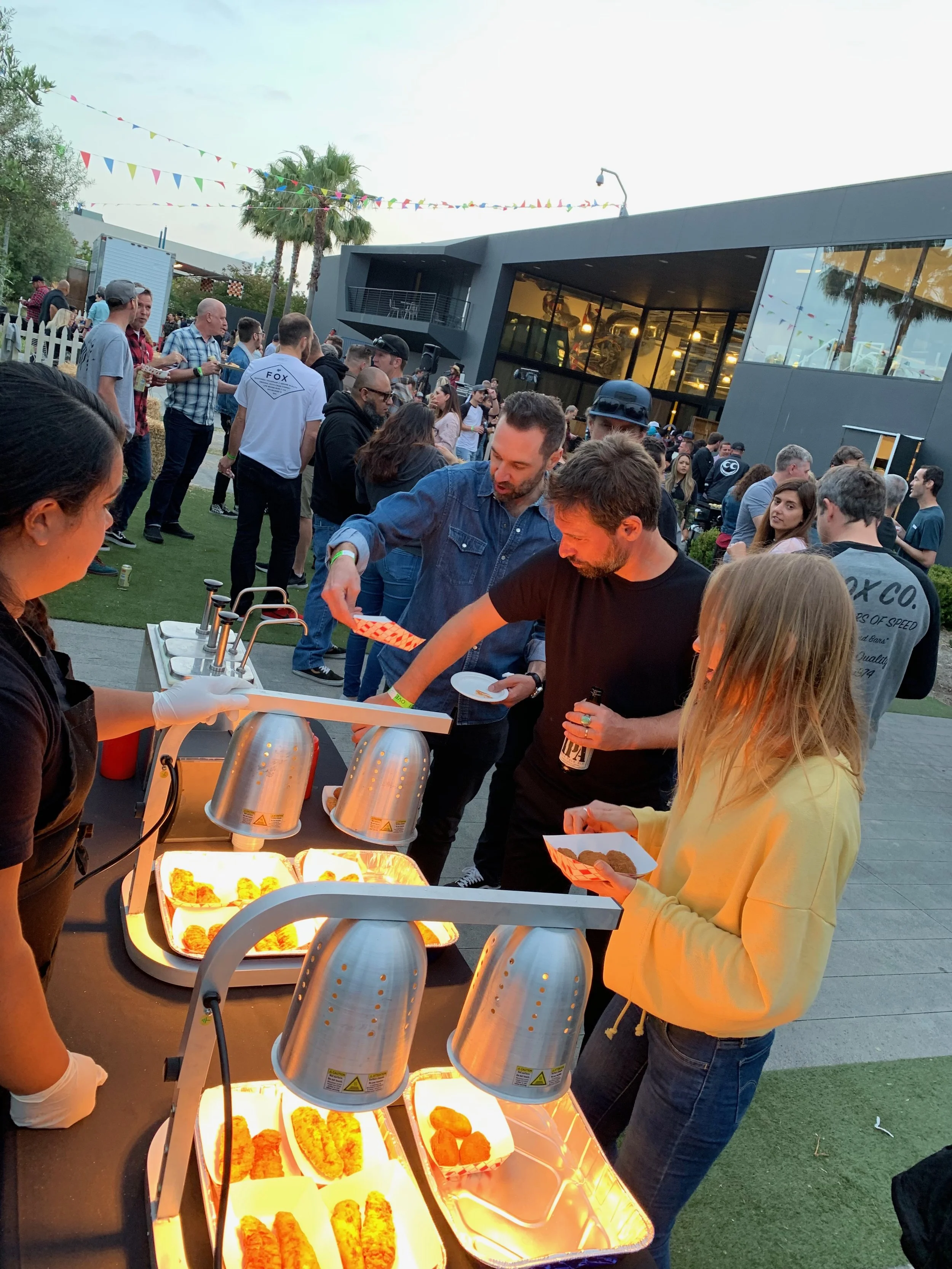 People at an outdoor event serving food from a chafing dish station with trays of hot foods, some individuals holding plates and serving utensils, set against a modern building with large glass windows, palm trees, and decorative bunting flags.