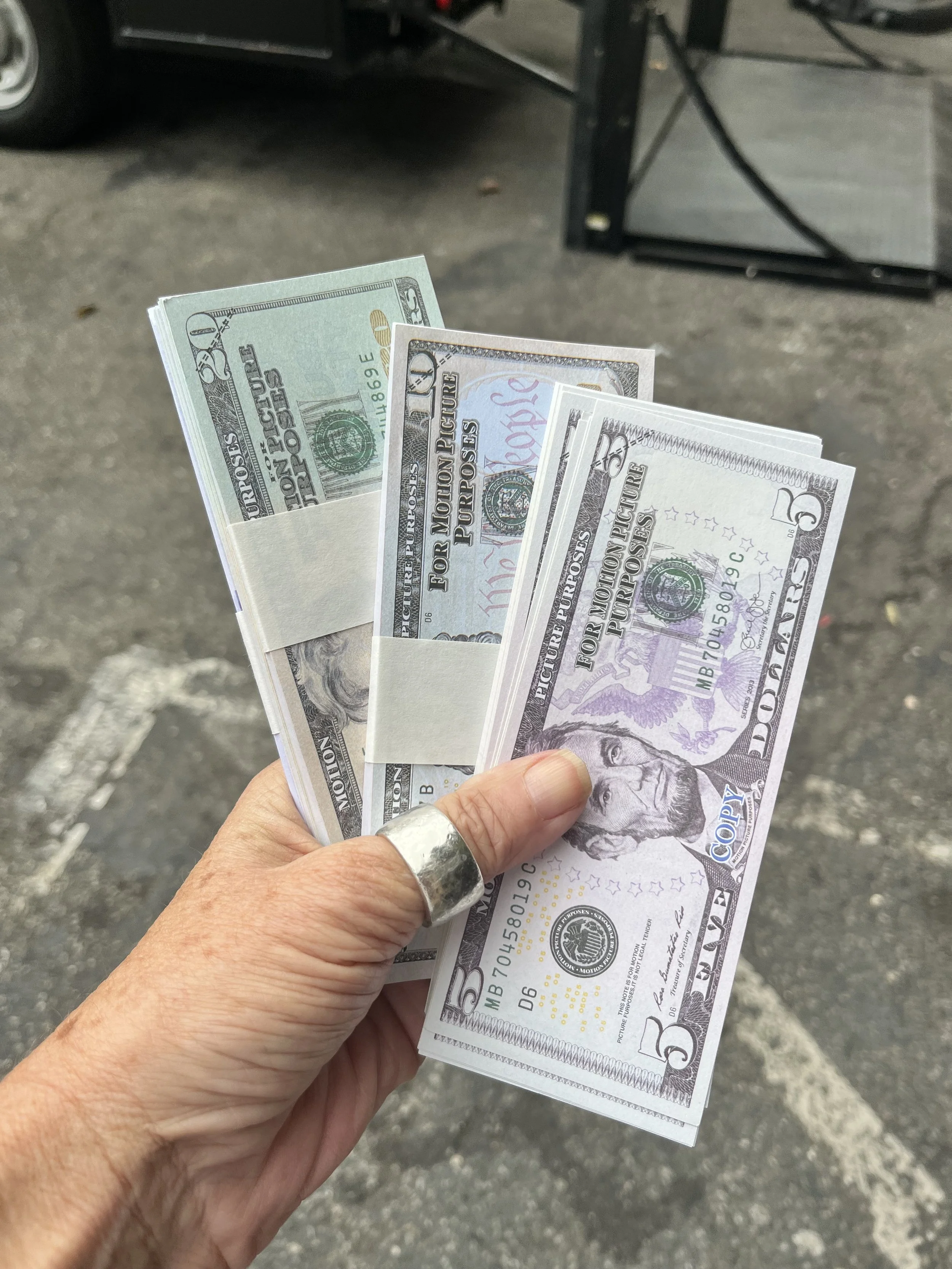 A person holding a small stack of $5 and $10 bills in front of a parking lot with a parked vehicle and a table or stand in the background.