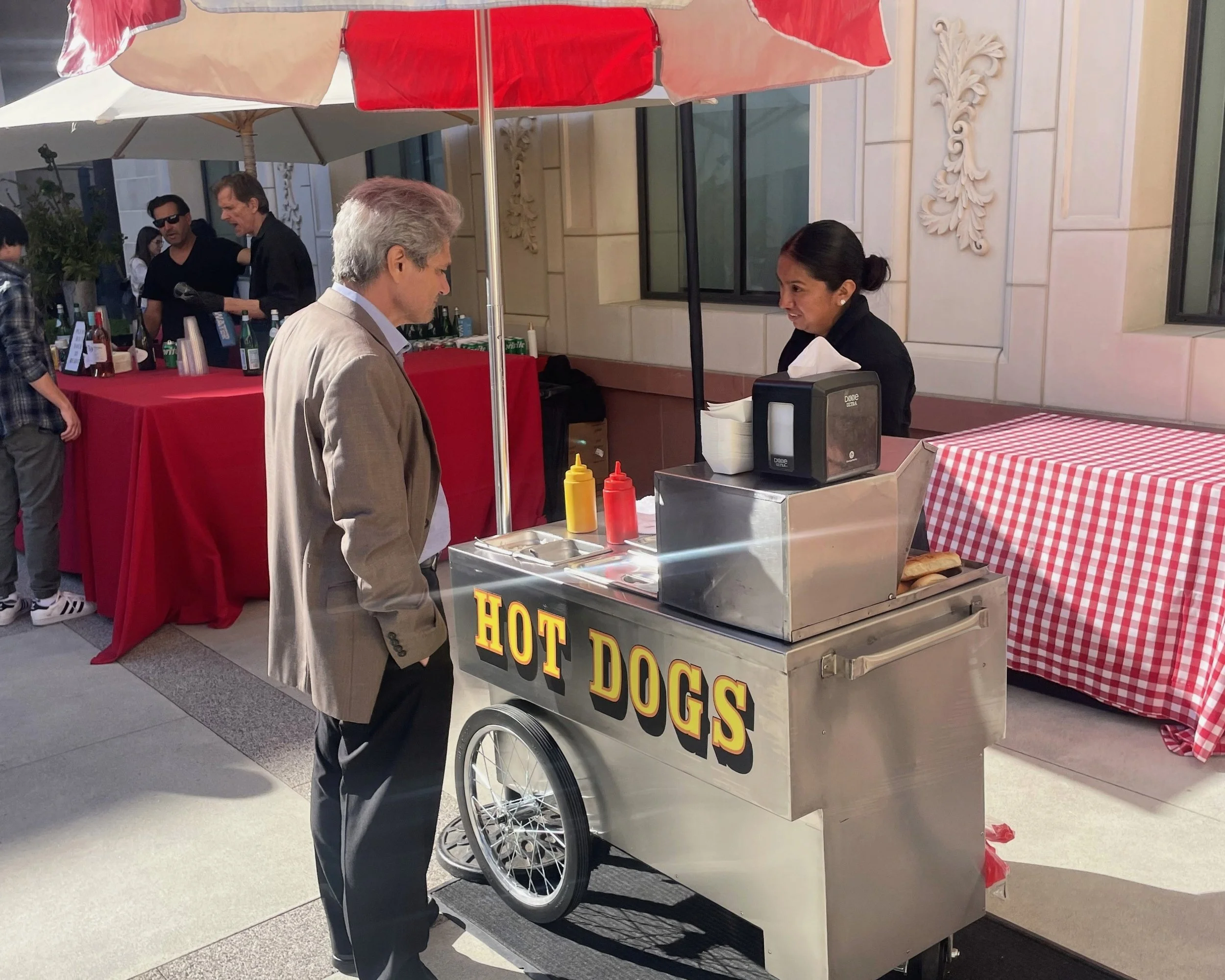 A man in a light-colored suit is ordering hot dogs from a street vendor cart that has a large sign reading 'HOT DOGS'. The cart has a red and white checkered tablecloth on a nearby counter and bottles of condiments on the kitchen counter. The vendor, a woman dressed in black, is preparing food behind the cart, which is outdoors on a city street with a cream-colored building in the background.