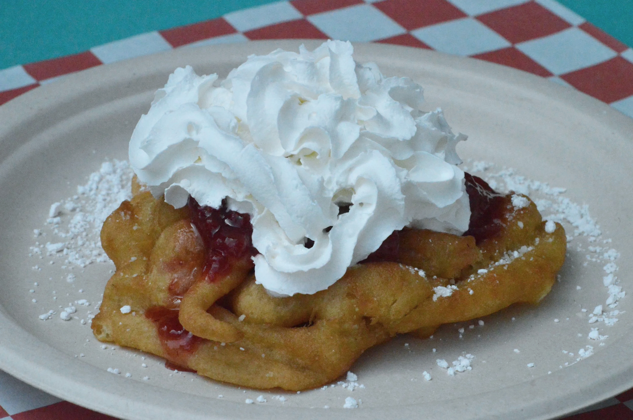 Funnel cake topped with whipped cream and berry filling on a paper plate, with powdered sugar around the base of the dessert.