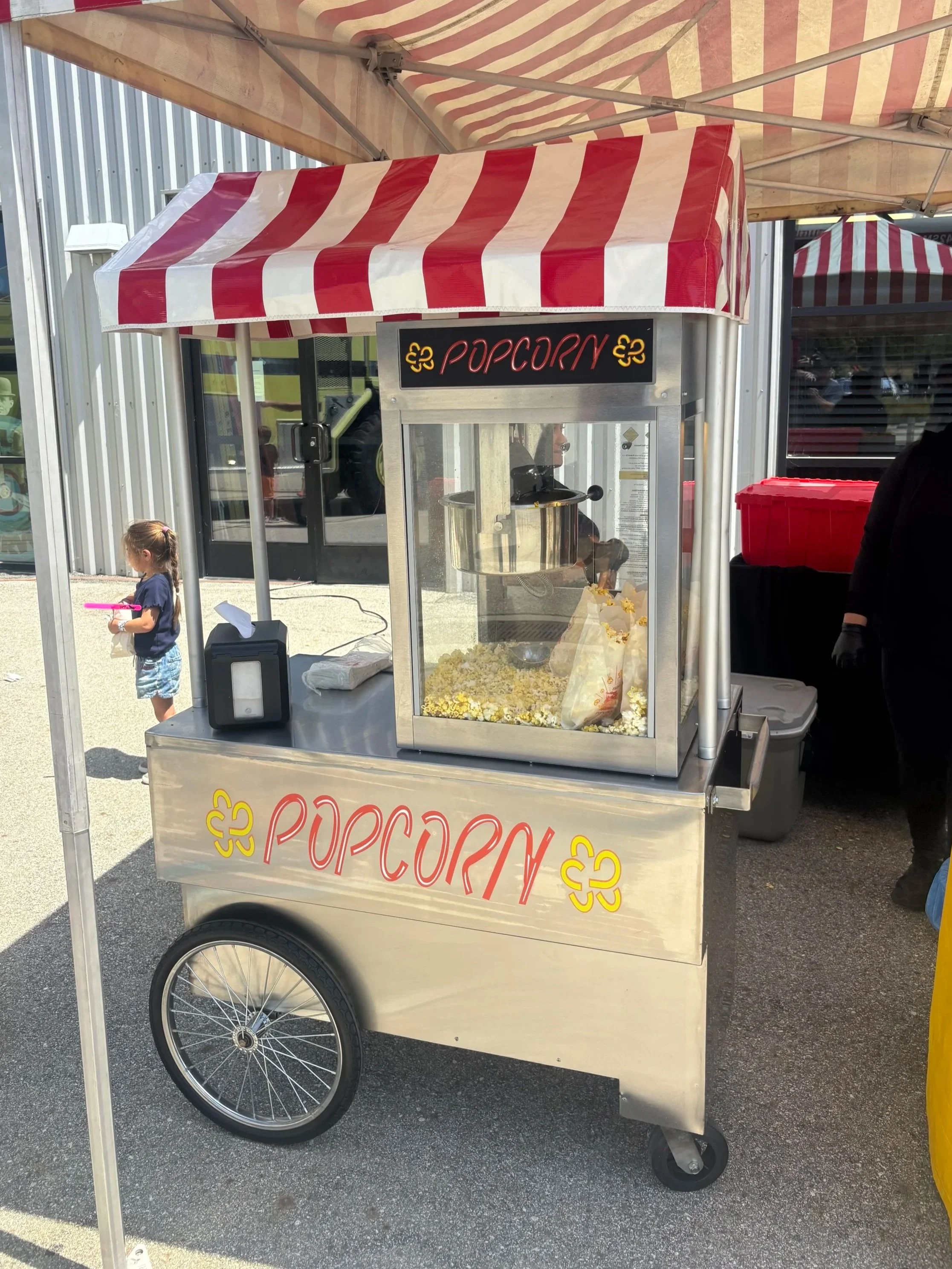 A popcorn cart with a red and white striped canopy, labeled 'Popcorn', selling fresh popcorn at an outdoor event. A young girl is standing nearby.