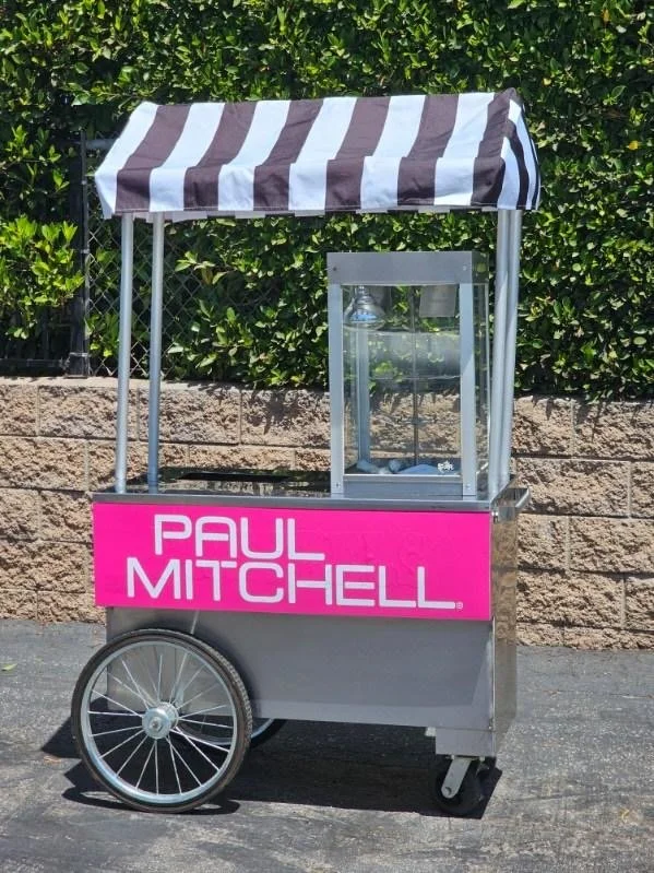 A mobile cart with a pink sign reading 'PAUL MITCHELL,' featuring a black and white striped canopy and large rear wheels, situated outdoors against a stone wall and green hedge.