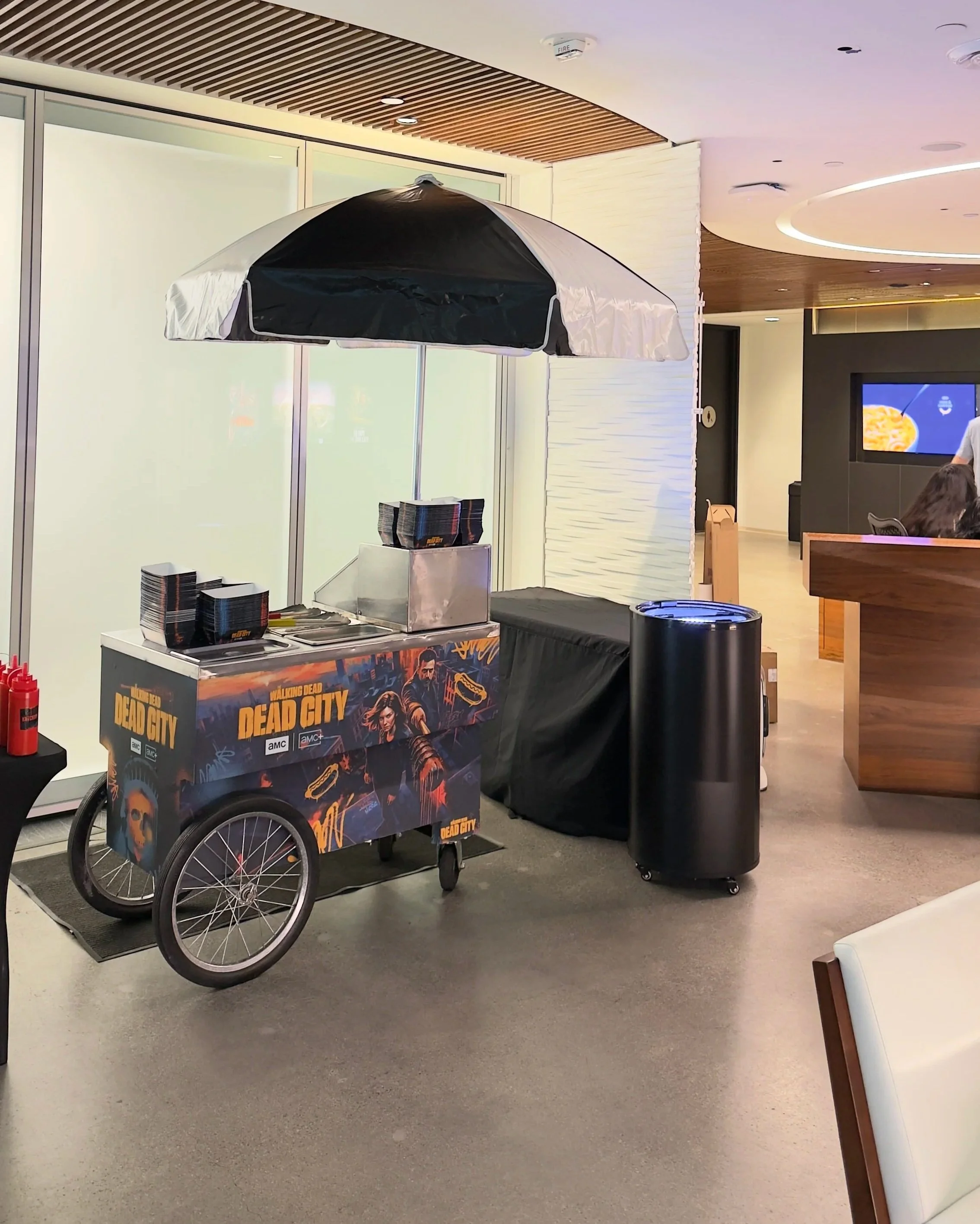 A food cart themed with the video game 'The Walking Dead Dead City,' featuring a black and orange design with characters from the game and a skull graphic. The cart is positioned indoors near a black trash can and a counter, with tables and chairs nearby in the background.