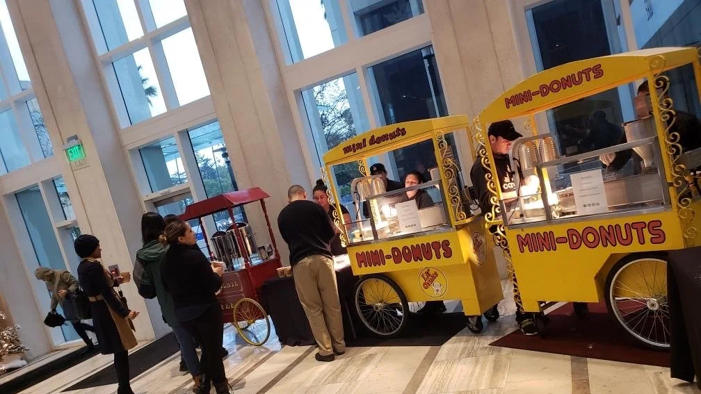People ordering mini donuts from yellow and red mini-donut carts inside a modern building with tall glass windows.