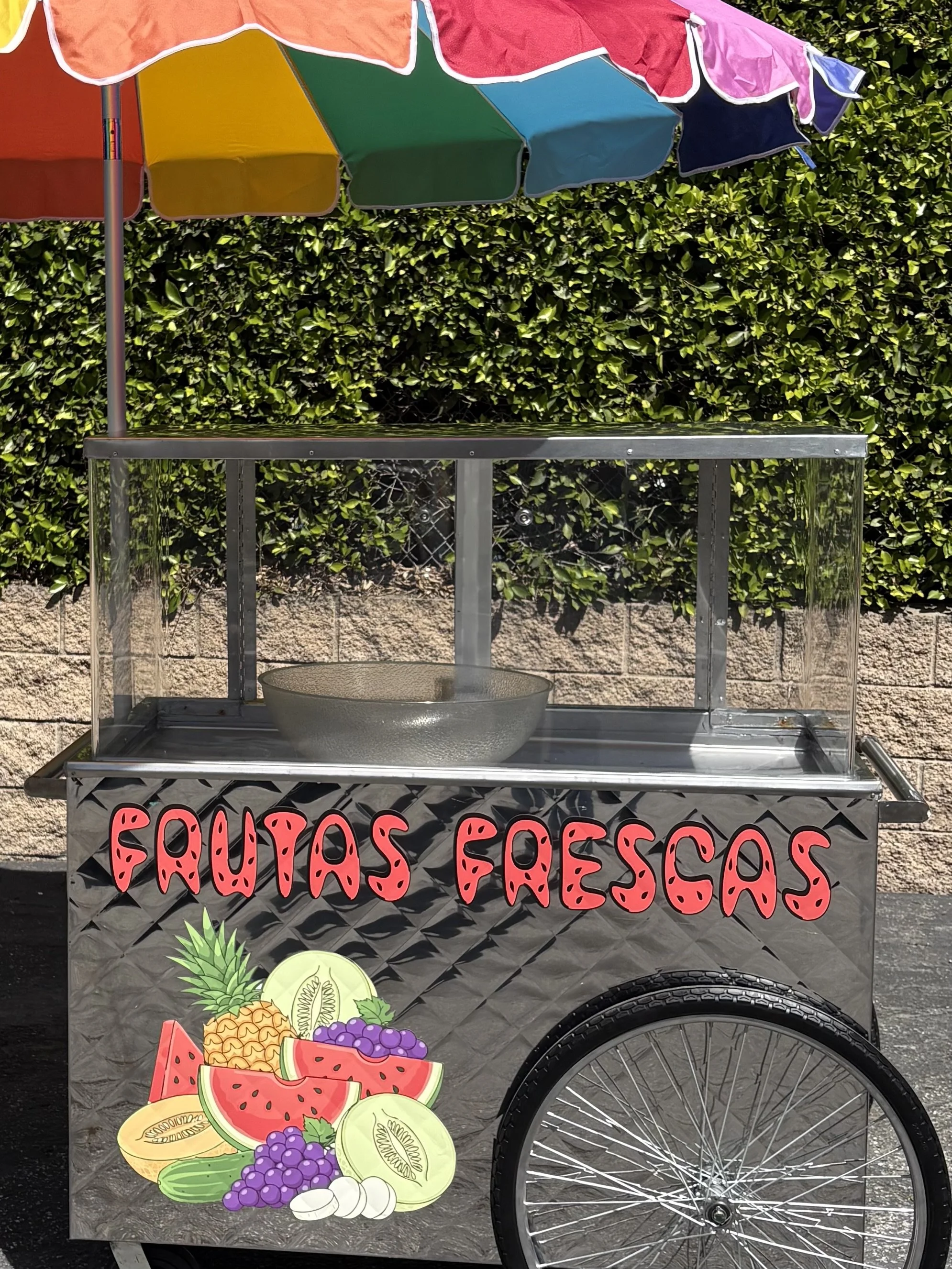 A fruit stand cart decorated with colorful fruit images and the words 'FRUTAS FRESCAS' in pink letters, featuring a large umbrella with multiple colors providing shade.