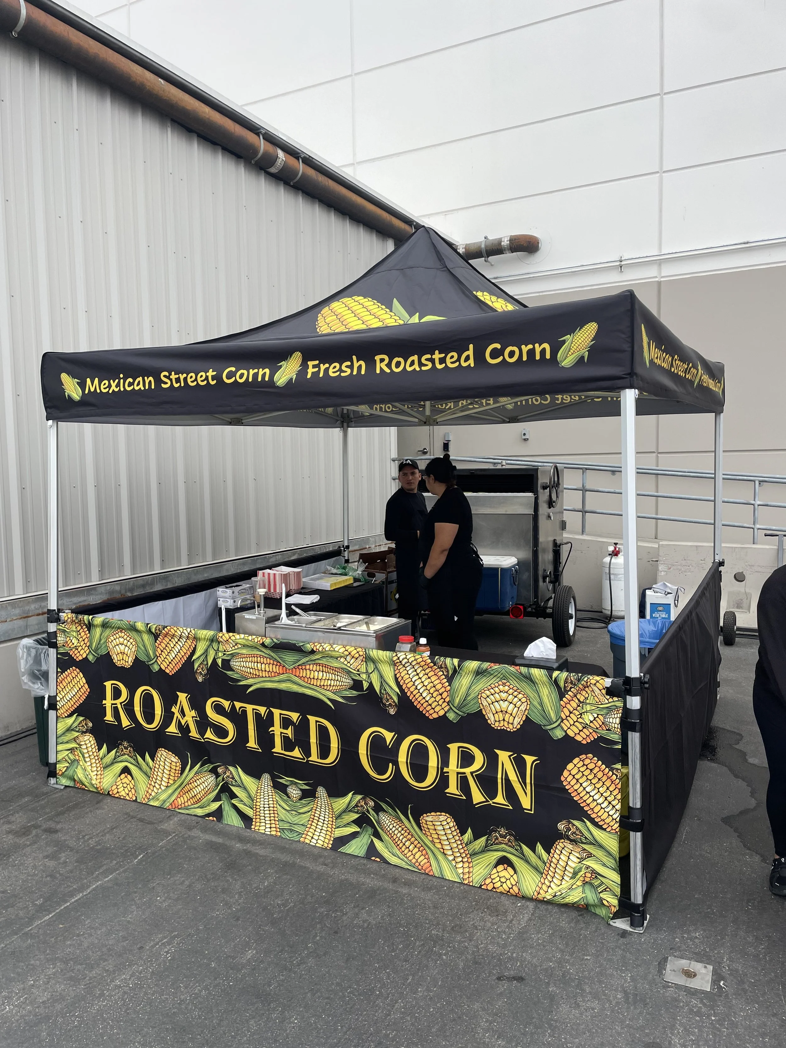 A street vendor stand with a black canopy that reads 'Mexican Street Corn' and 'Fresh Roasted Corn' with illustrations of corn on the cob. Two people are behind the stand preparing food, with supplies and a grill visible in the setup.