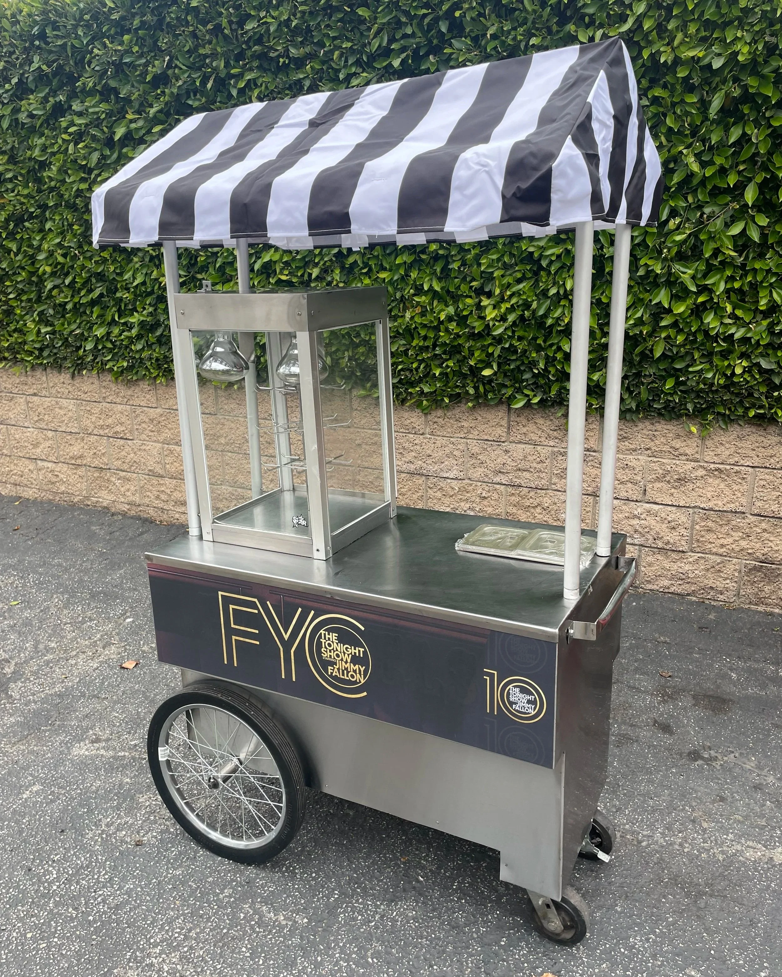 A carnival style food cart with a striped black and white canopy, featuring branding for The Tonight Show starring Jimmy Fallon, parked on asphalt with a brick wall and green hedge in the background.