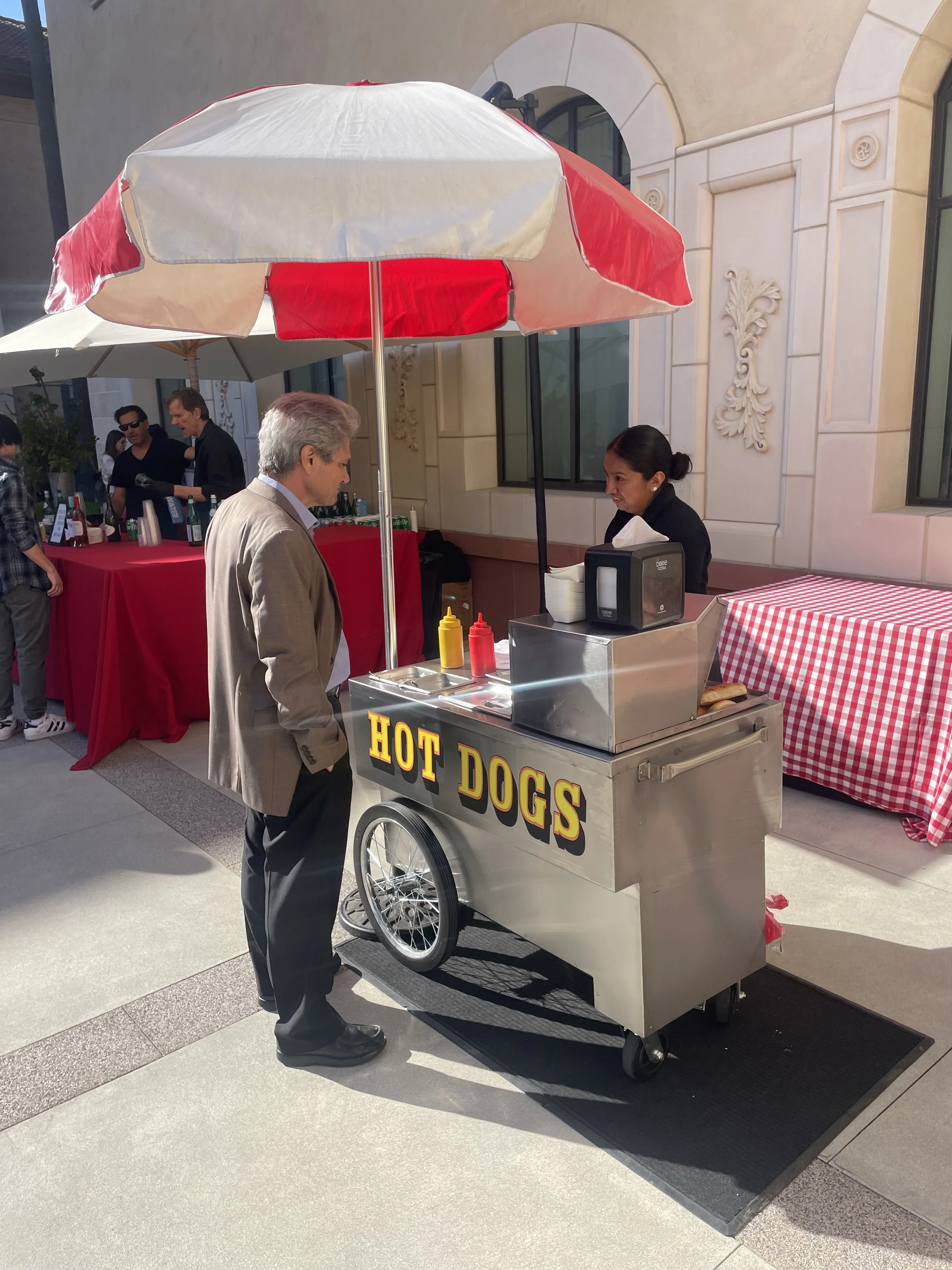 A man orders food from a hot dog stand with a large red and white umbrella. The stand has a sign that reads 'HOT DOGS' and condiments are available on the cart. The scene takes place outdoors in front of a building with decorative architectural details.