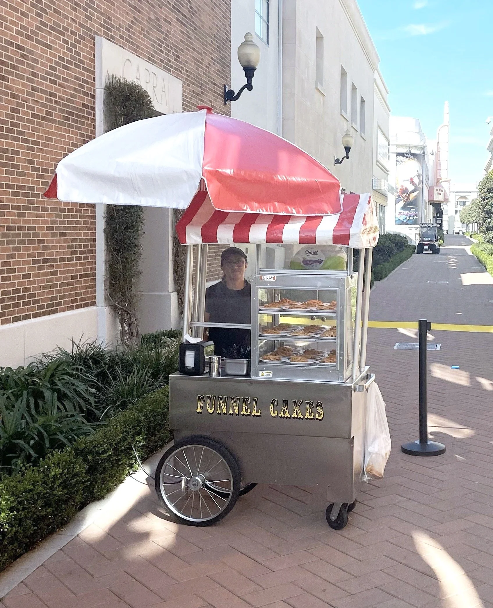 A street food cart selling funnel cakes with a striped red and white canopy, located on a sidewalk with brick and white building facades, next to greenery and a lamppost.