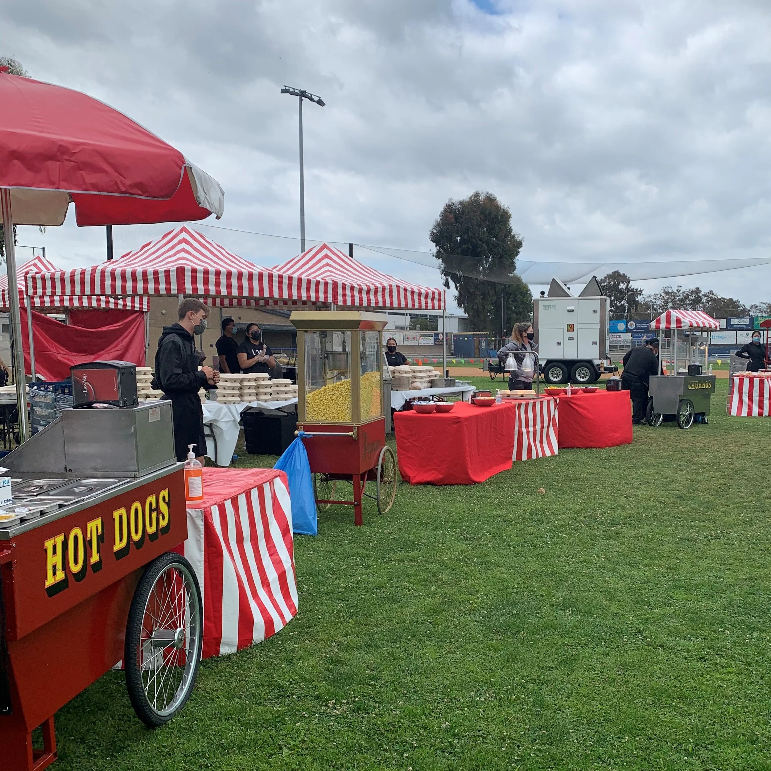 Outdoor setup with red and white striped tents and a red food cart with a sign for hot dogs, people wearing masks, cloudy sky, and trees in the background.