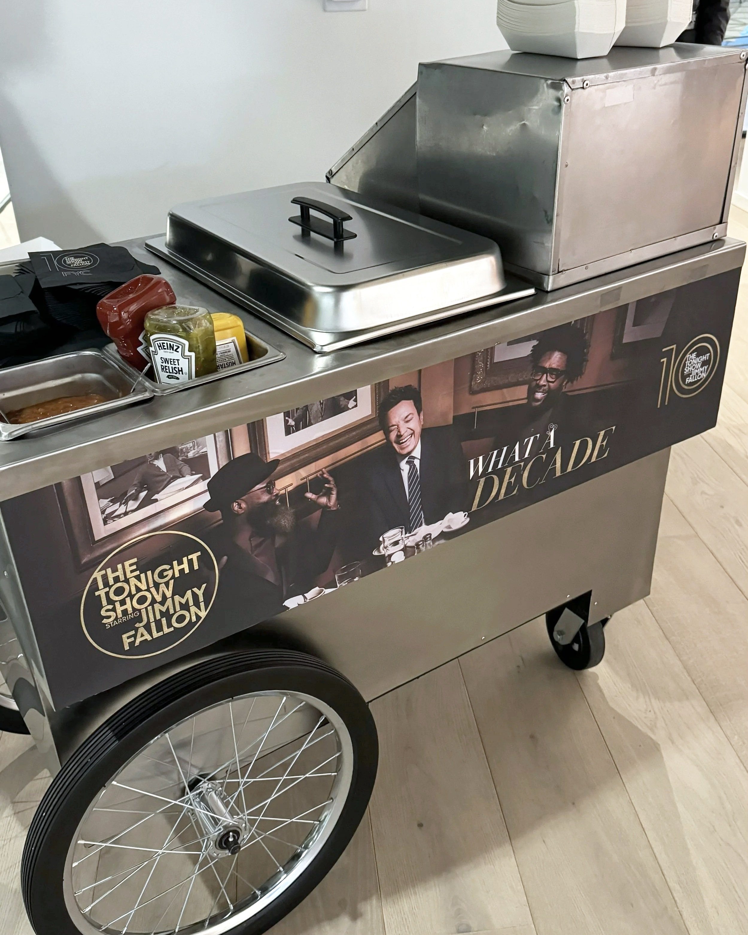 A hotdog cart with a poster featuring comedians from The Tonight Show starring Jimmy Fallon, and a food service cart with condiments and food compartments at an indoor location.