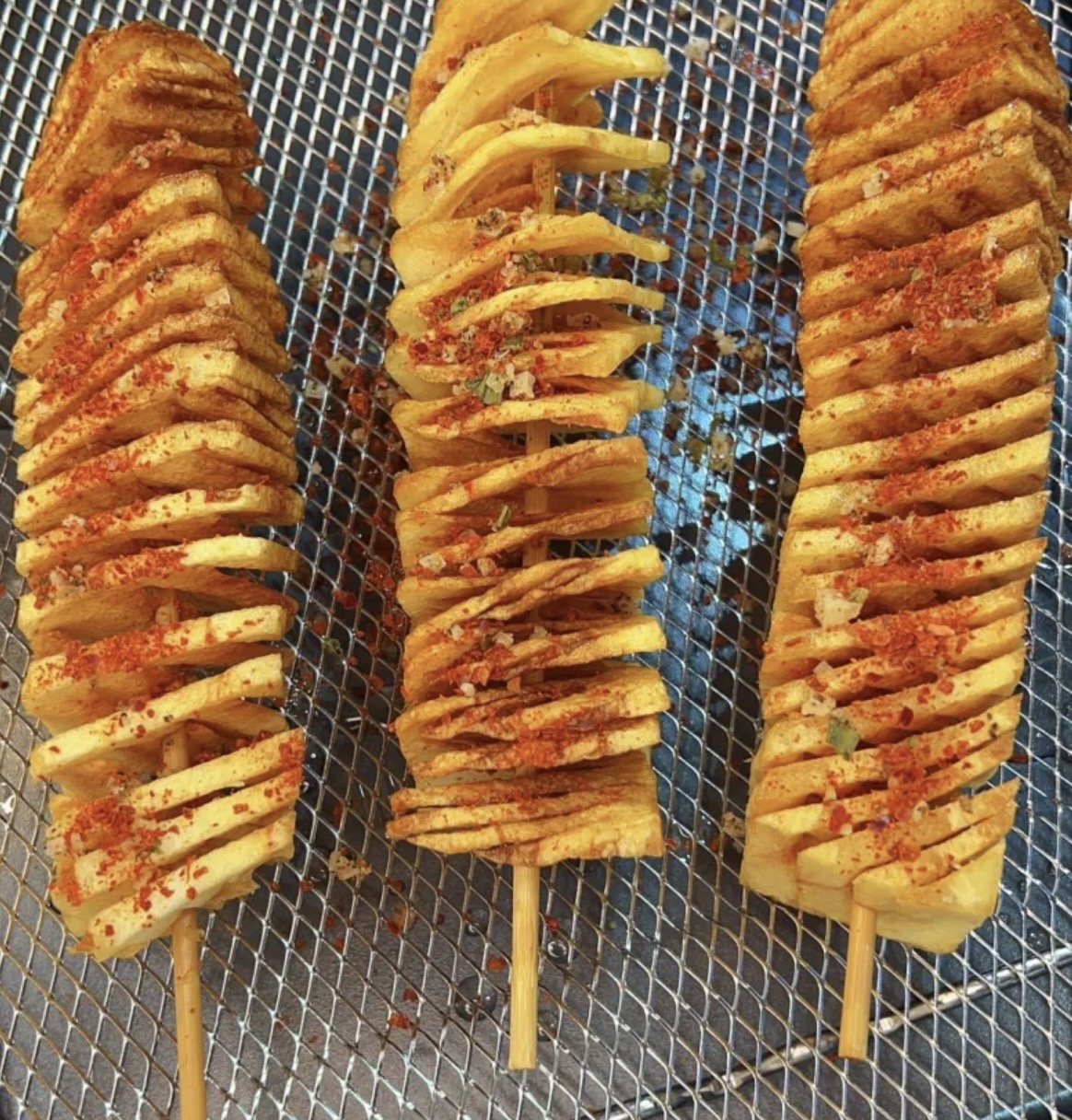 Three skewers of spiral-cut potatoes seasoned with red spice, resting on a metal grilling rack.