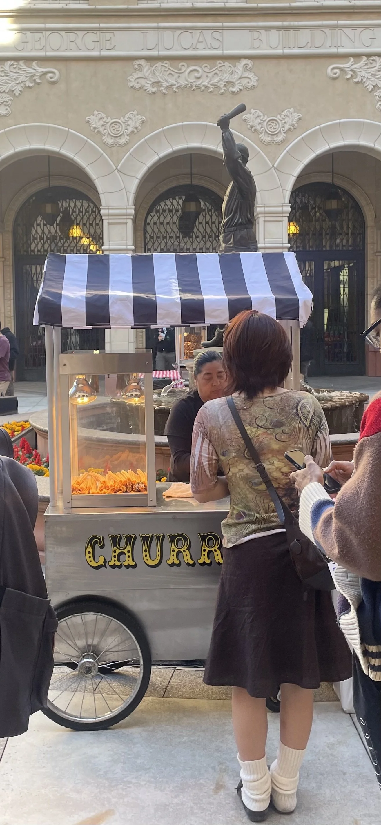 A street scene featuring a churro stand with a black and white striped awning, people gathered around, and a fountain with a statue in the background at the George Lucas Building.