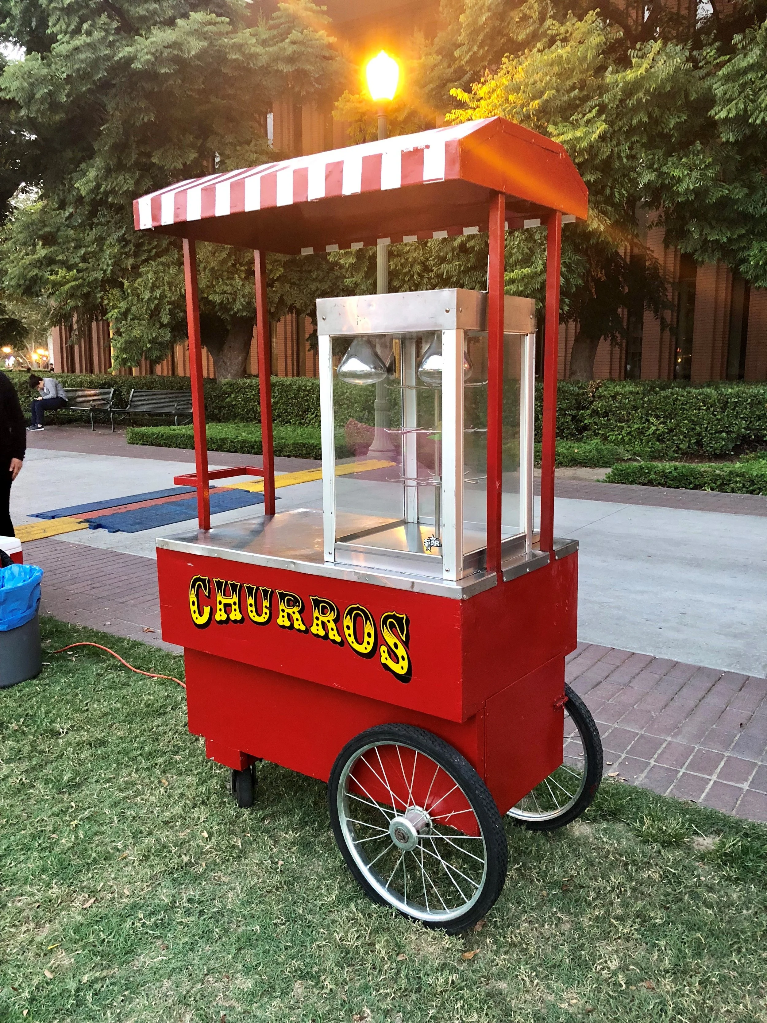 A red jalapeno popper stand with the word 'CHURROS' written on it in yellow and black letters, set in a park with trees and benches, and a man sitting on a bench in the background.