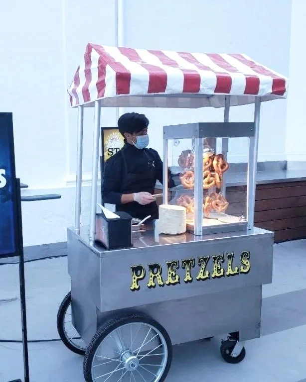 A person wearing a face mask and black chef's jacket preparing pretzels at a mobile pretzel stand with a red and white striped canopy.