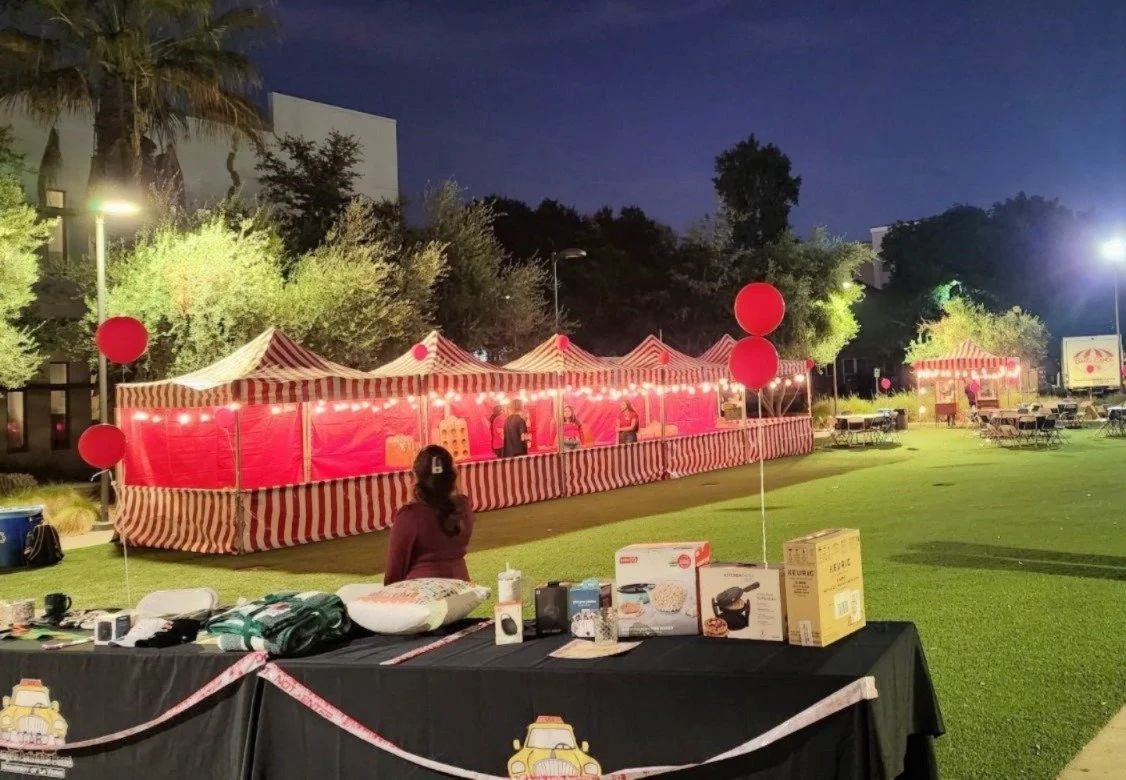 Outdoor carnival or fair setup at night with red and white striped tents, red balloons, and tables with prizes or items, illuminated by bright lights, with a grassy area and trees in the background.