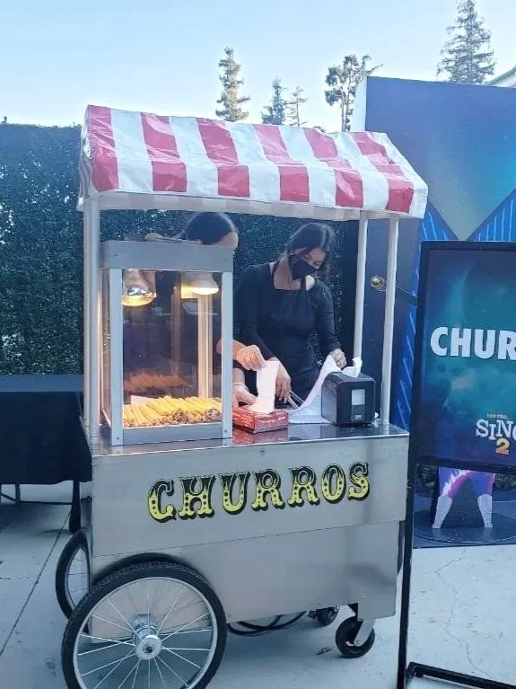A street food cart with a red and white striped canopy selling churros, with two women preparing the food in an outdoor setting.