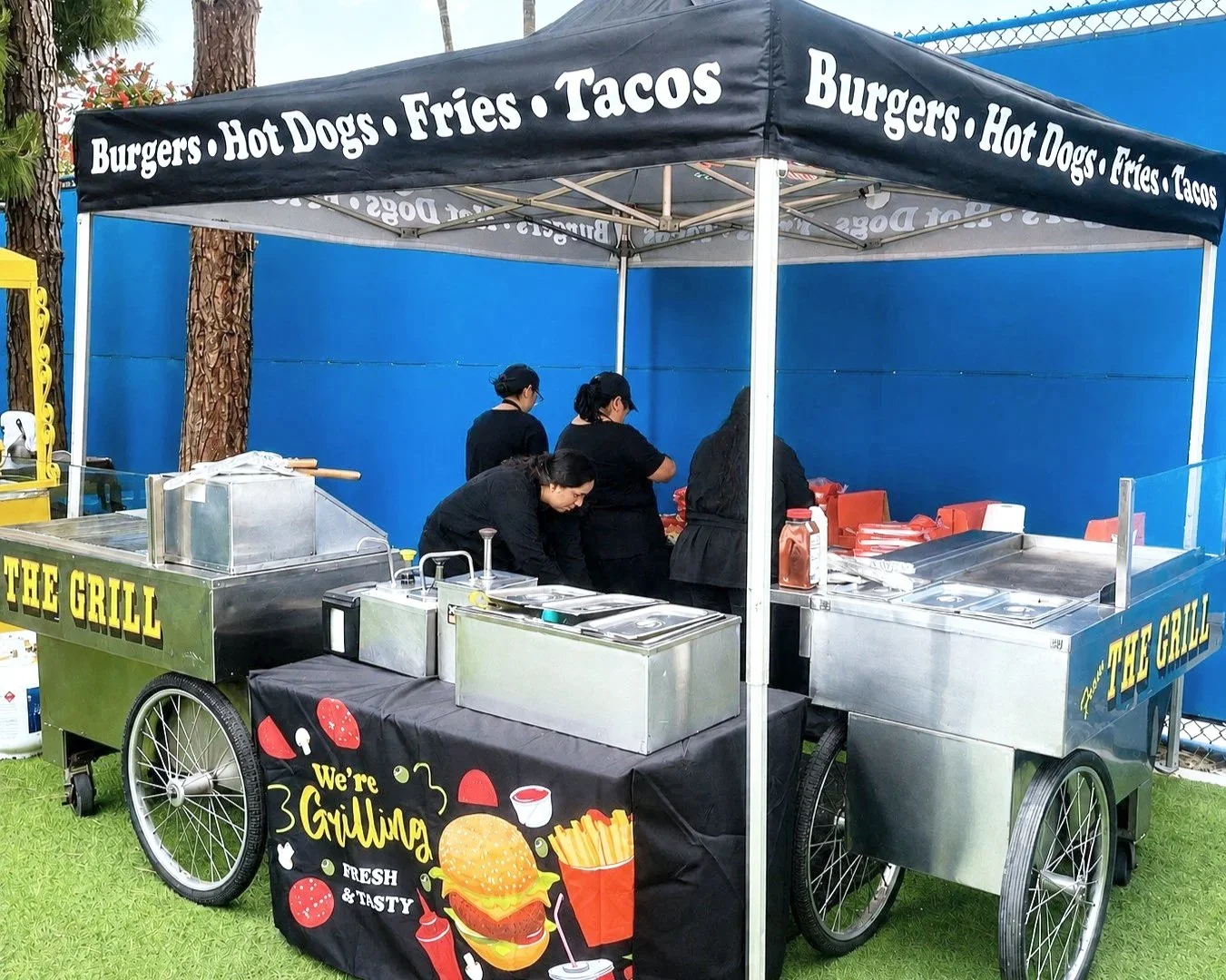 Food cart with a black canopy advertising burgers, hot dogs, fries, and tacos; four people working behind the cart, with a sign saying "We're Grilling, Fresh & Tasty", featuring a burger, fries, and sauce illustrations.