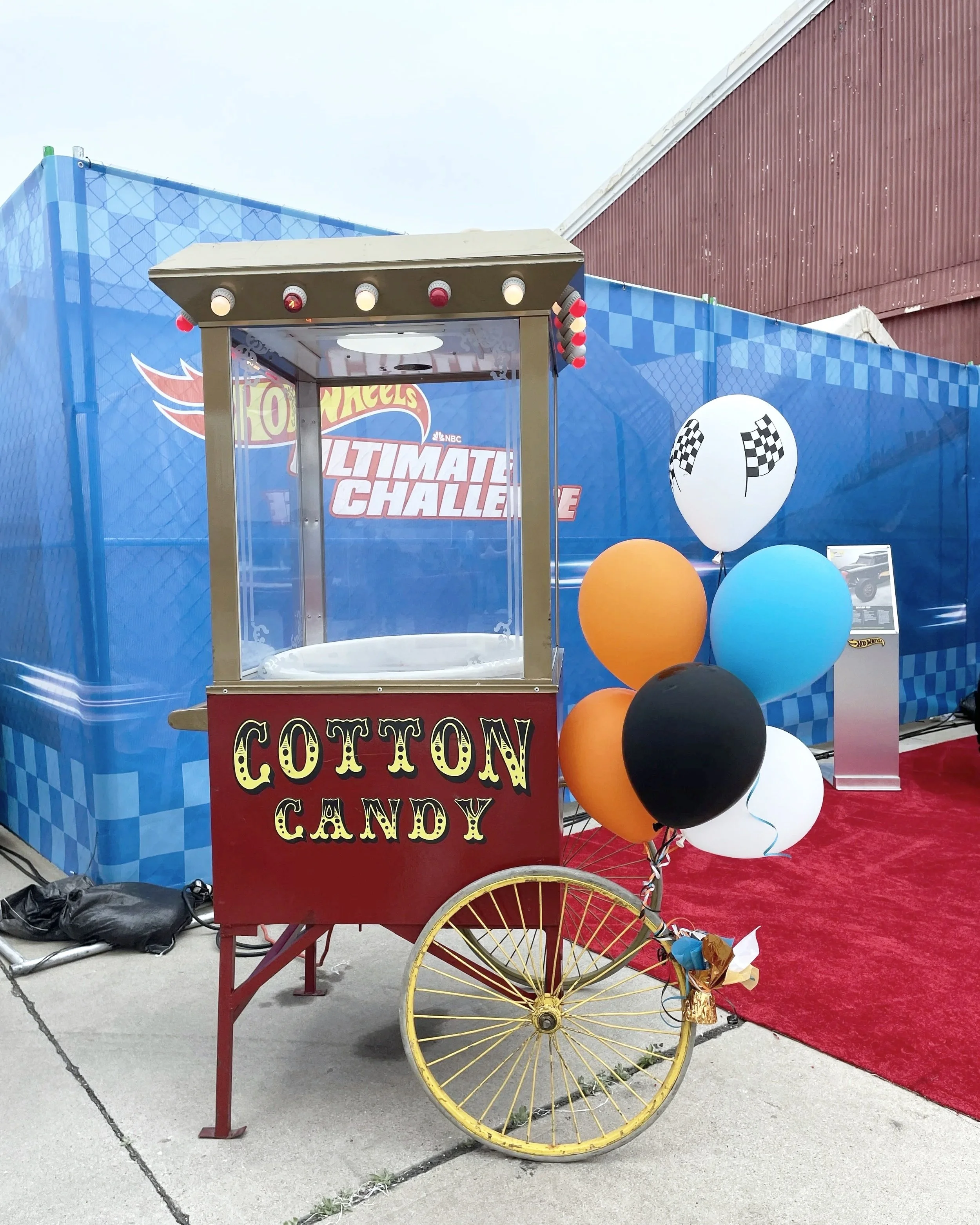 A cotton candy cart with a colorful red sign, decorated with balloons, set up on a sidewalk in front of a blue booth with checkered pattern at an event.