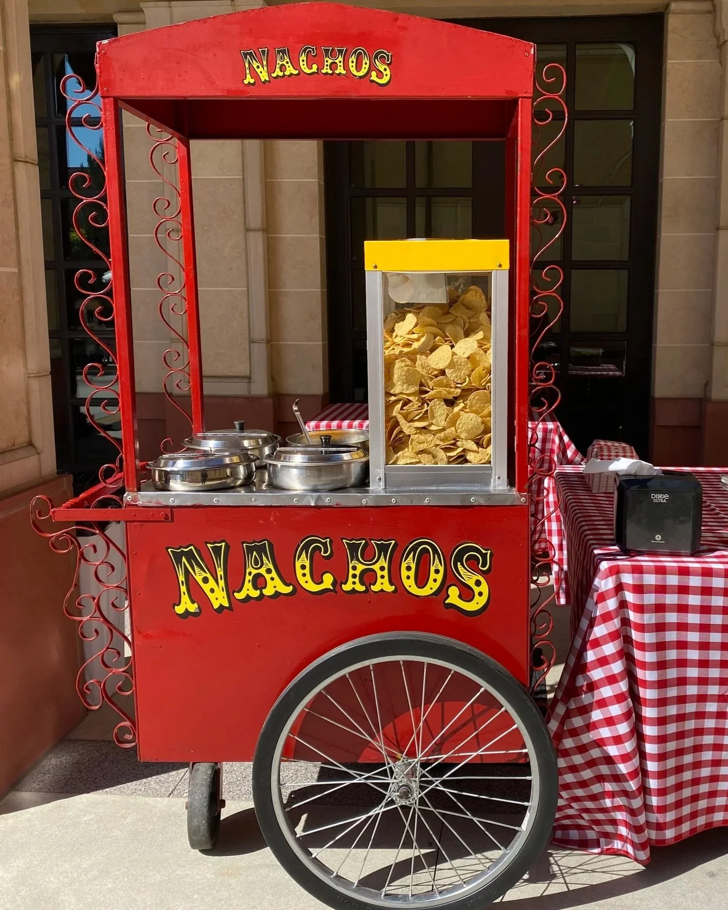A red nacho stand on wheels with a yellow sign, displaying a container of potato chips, two metal chafing dishes, and a table with a red and white checkered tablecloth.