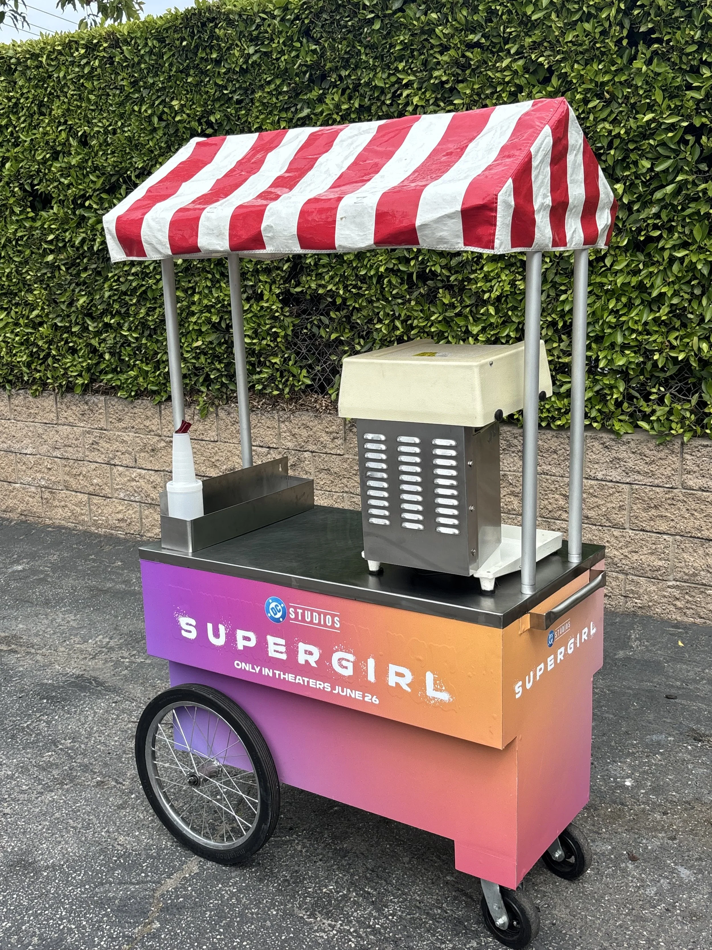 A mobile food cart with a red and white striped canopy promoting the movie Supergirl, featuring the DC Studios logo and release date June 26, parked on a paved surface against a background of green bushes.