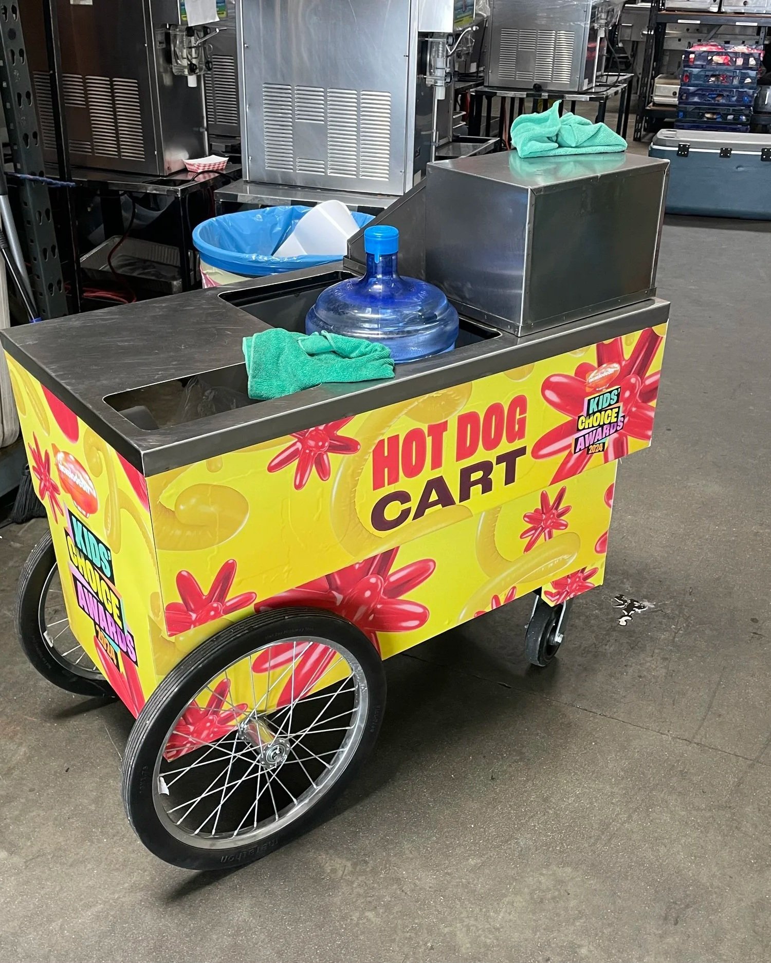 A colorful kids' hot dog cart with wheels, decorated with red star shapes on a yellow background. There is a person kneeling in front of the cart adding special branding for a spcial event..