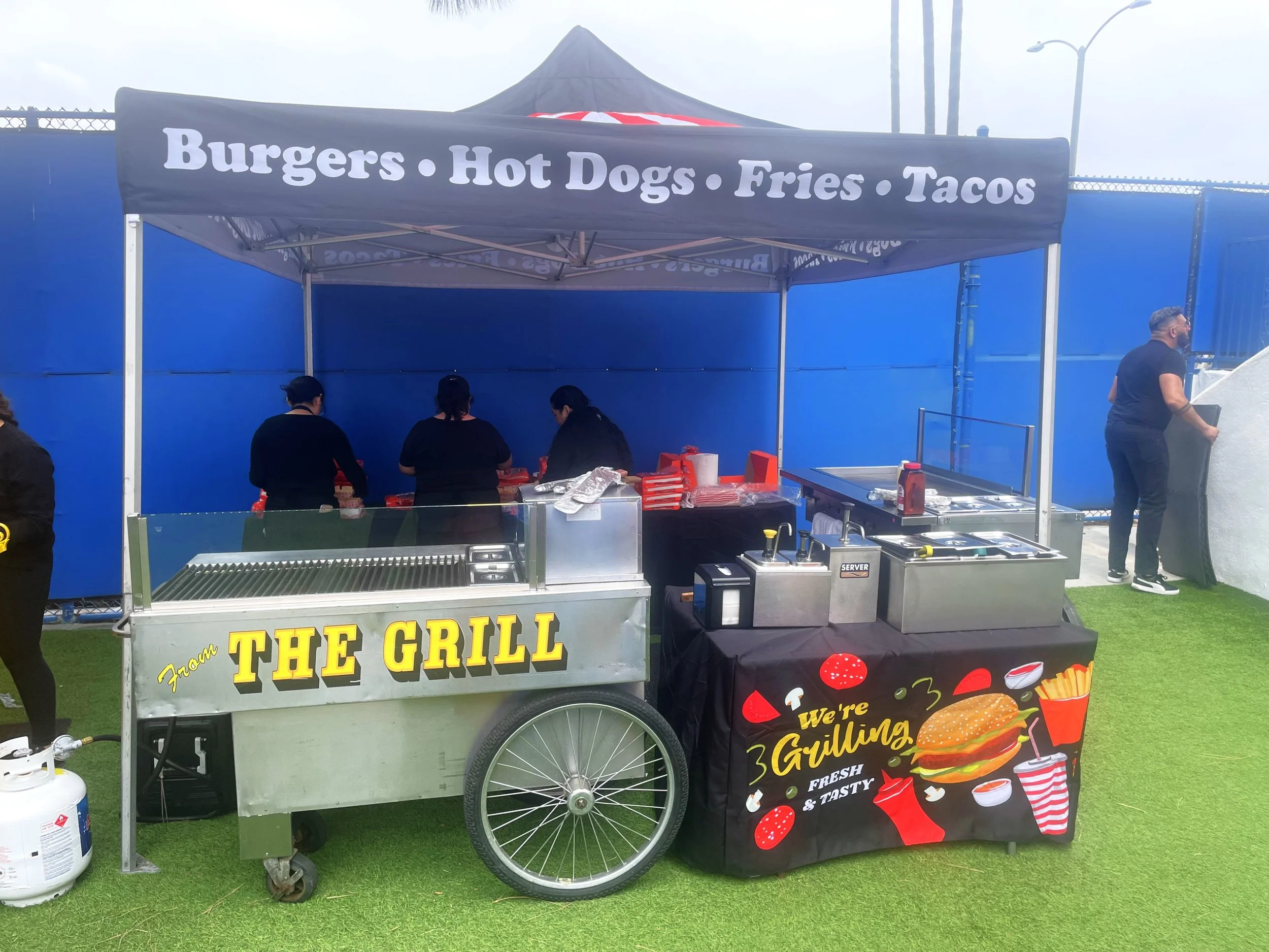 A food cart with a sign advertising burgers, hot dogs, fries, and tacos. The cart is black with colorful food advertisements and is set up on a grassy area. There are people preparing food inside the cart, with a man standing on the right side.