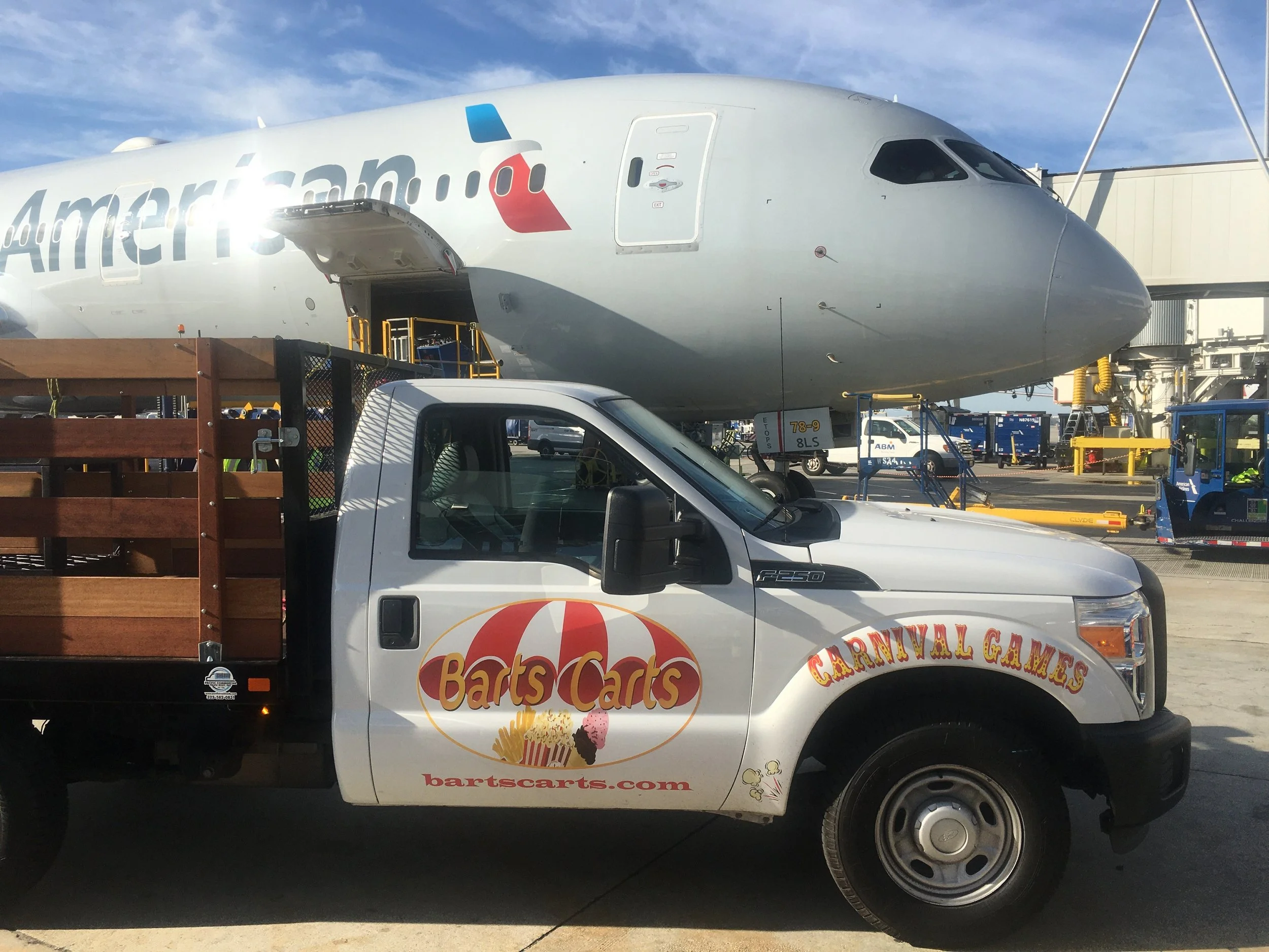 A white truck with a dessert shop logo, parked at an outdoor carnival games area. In the background, there is a large airplane and some carnival equipment.