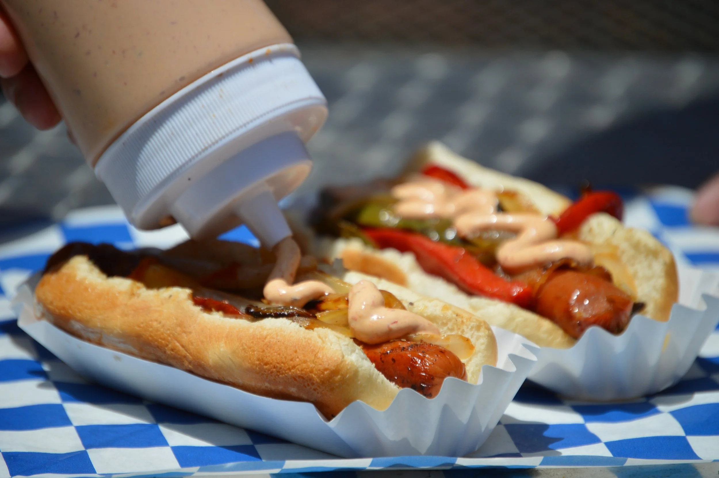 Close-up of a hot dog in a bun topped with ketchup, mustard, relish, and grilled onions, with a bottle of creamy sauce being squeezed onto it, placed on a blue and white checkered paper.