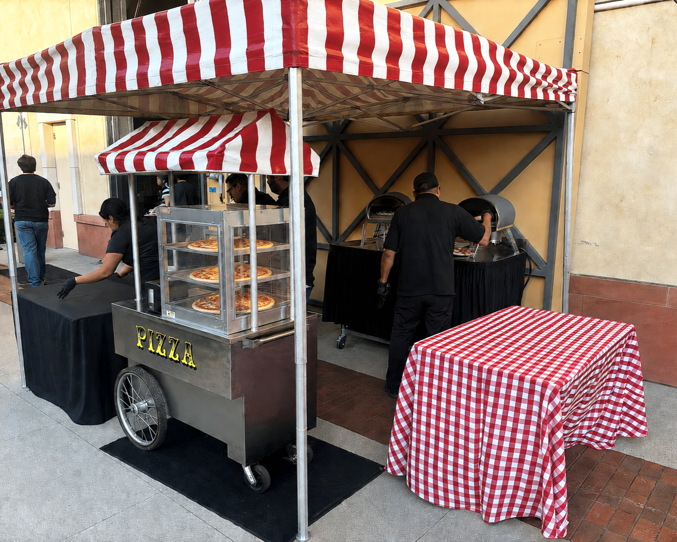 A pizza stand with a black table labeled 'PIZZA', a cart with pizza slices on display, and a red-and-white checkered canopy. Two workers in black are preparing food at the stand, and there is an empty checkered tablecloth-covered table in front.