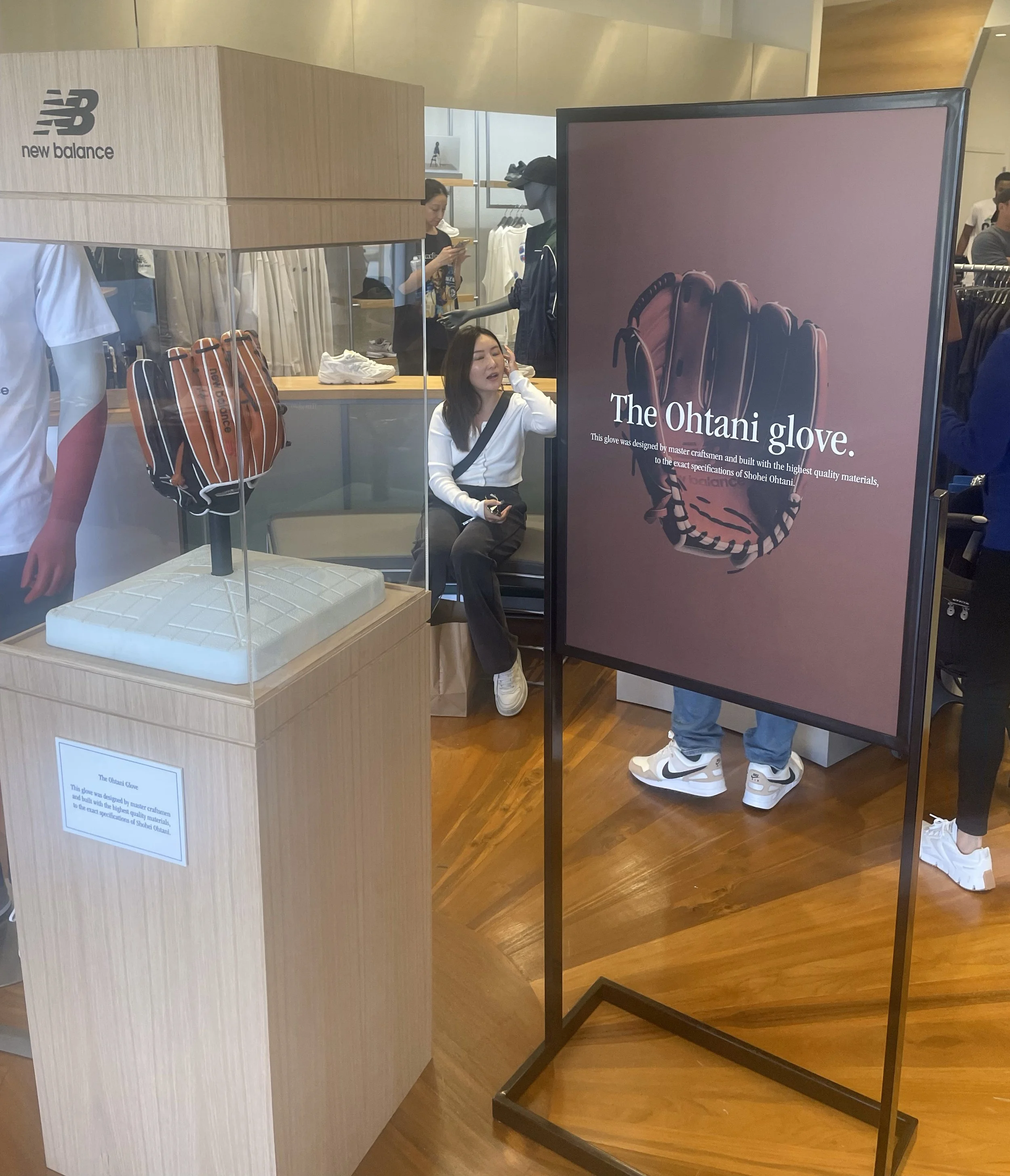 A retail store displays a New Balance backpack, a mannequin partially visible wearing white and red sportswear, and promotional signage for the 'Ohtani Glove,' describing it as designed by master craftsmen with high-quality materials, on a wooden flo