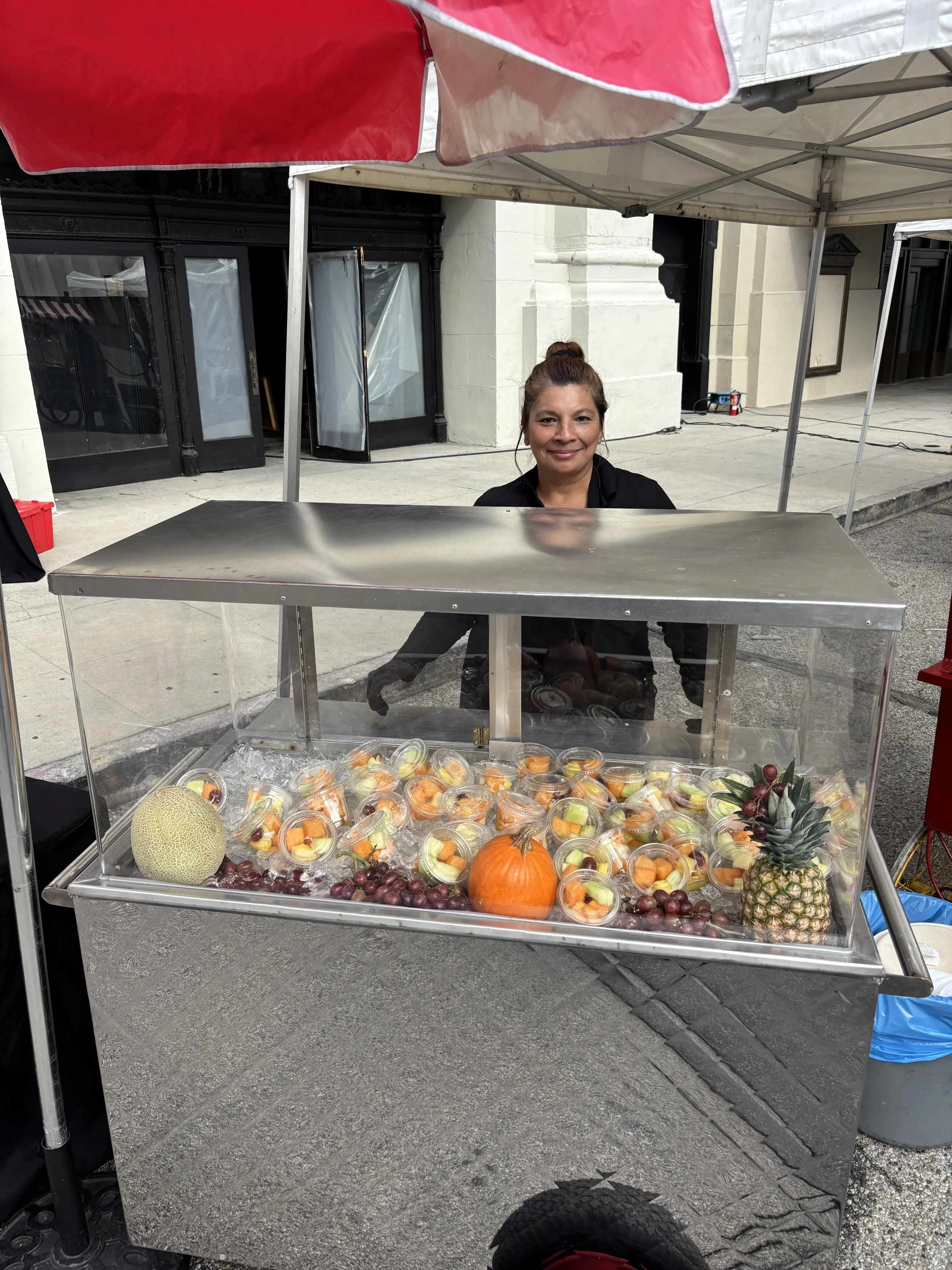 A woman is standing behind a food cart on a city sidewalk, with a display of pre-cut fruits like melon, grapes, pineapple, and cantaloupe in plastic cups. The cart is covered with a glass display case, and there is a pineapple and a cantaloupe on the counter. She is smiling and wearing a black shirt. The background shows a building with black and white doors and a concrete sidewalk, and there is a red and white canopy above her.