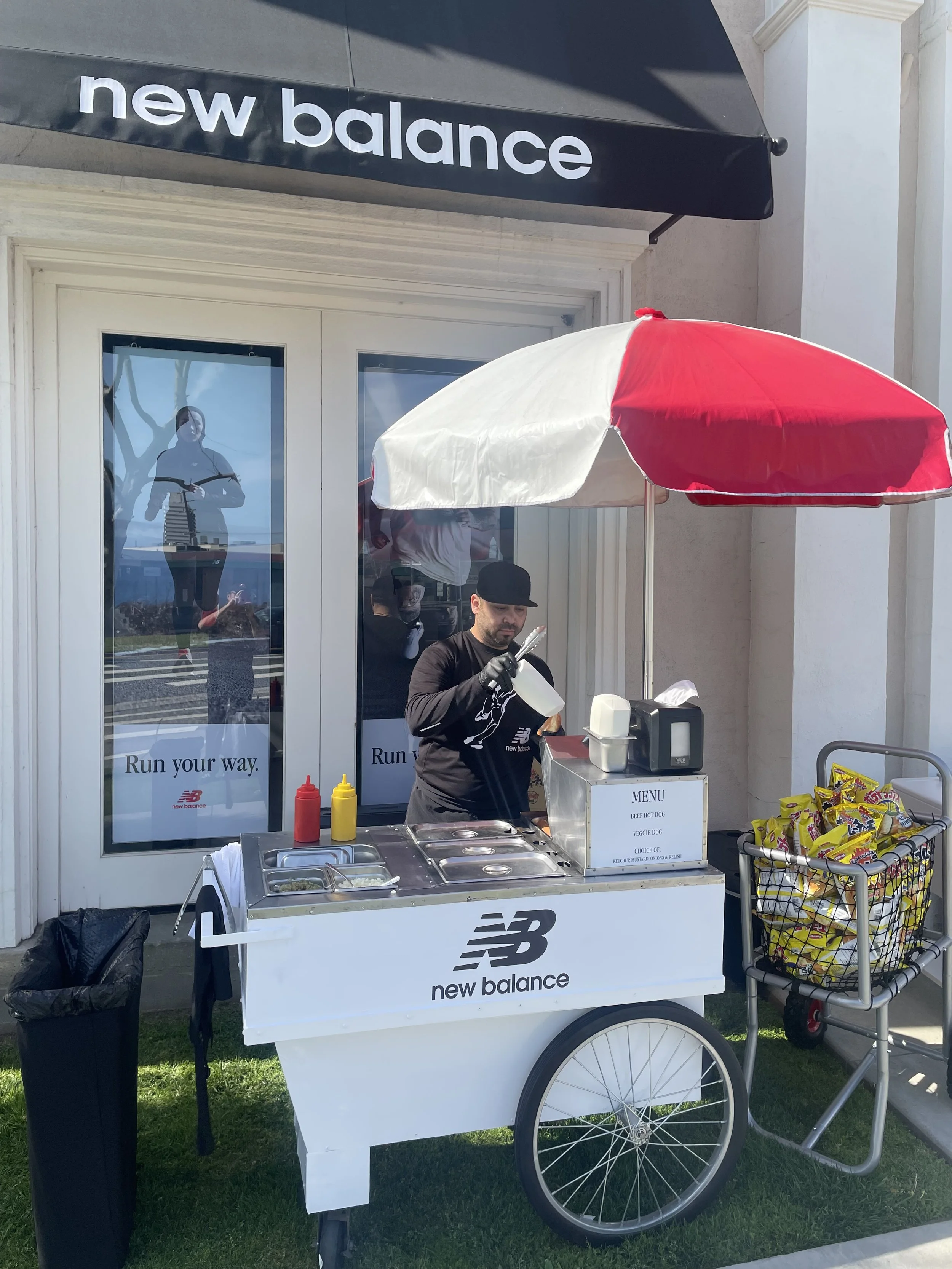 A man working at a New Balance food cart with a red and white umbrella, beside a cart filled with snack chips, and behind a glass door with running advertisements.