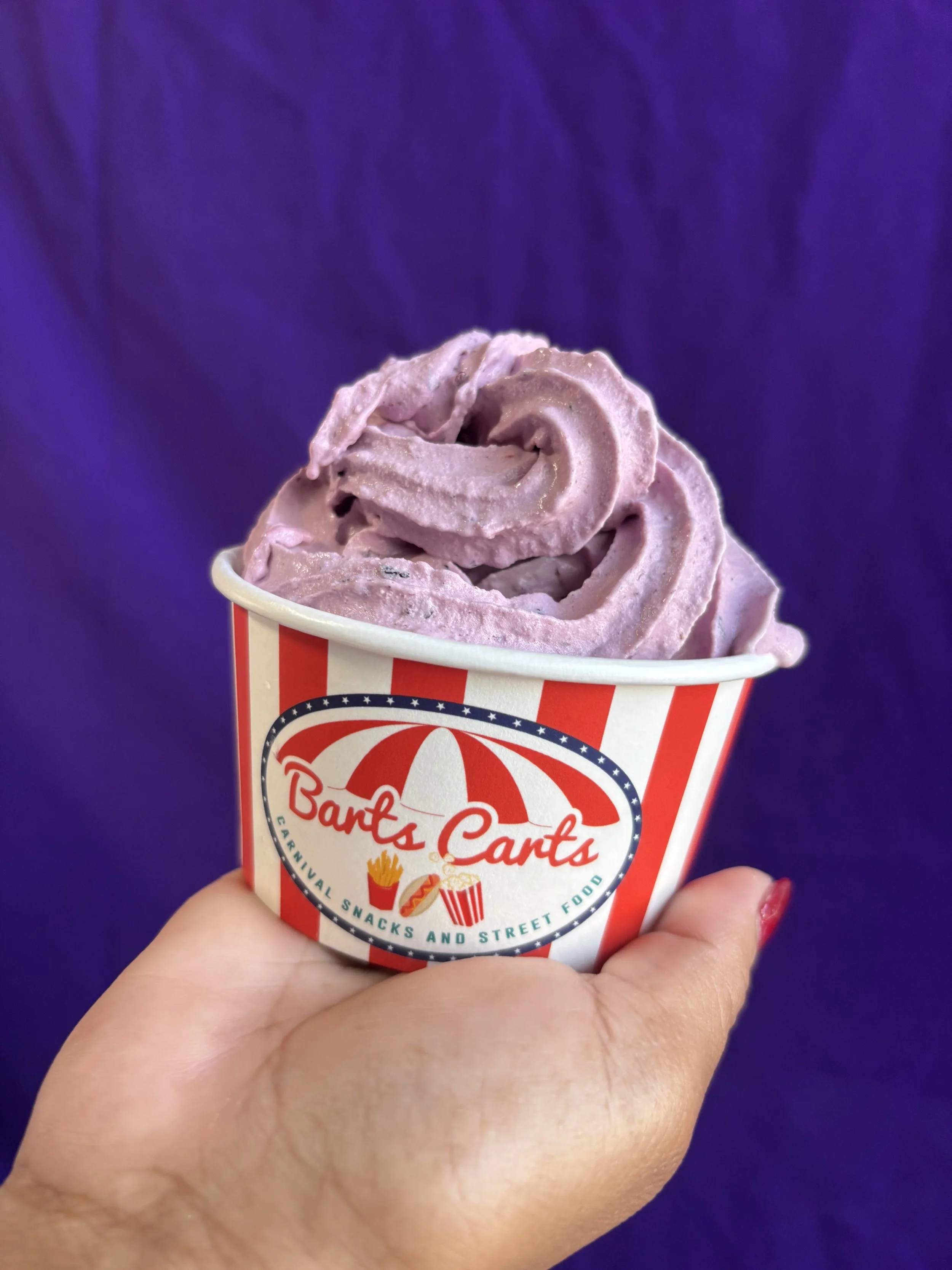 A hand holding a cup of purple soft-serve ice cream labeled Barf's Carts, with a background of dark purple draped fabric.