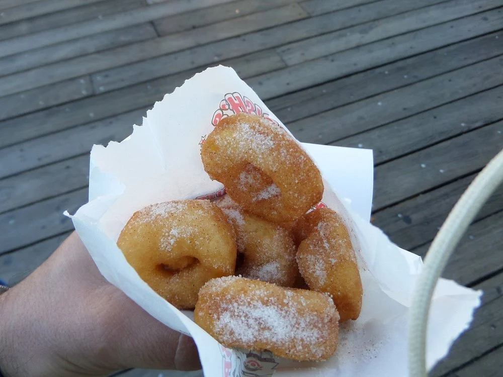A hand holding a paper cone filled with four fried apple fritters dusted with powdered sugar, against a wooden deck background.