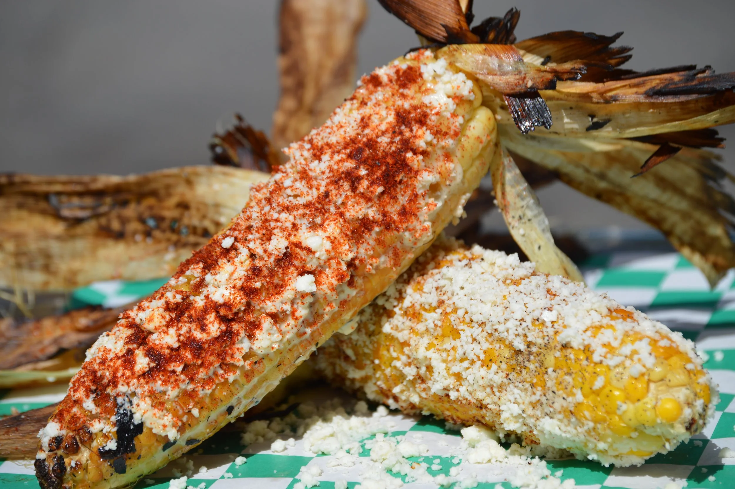 Close-up of Mexican street corn on a green and white checkered paper, topped with chili powder, cheese, and spices, with some corn kernels spilled on the paper.