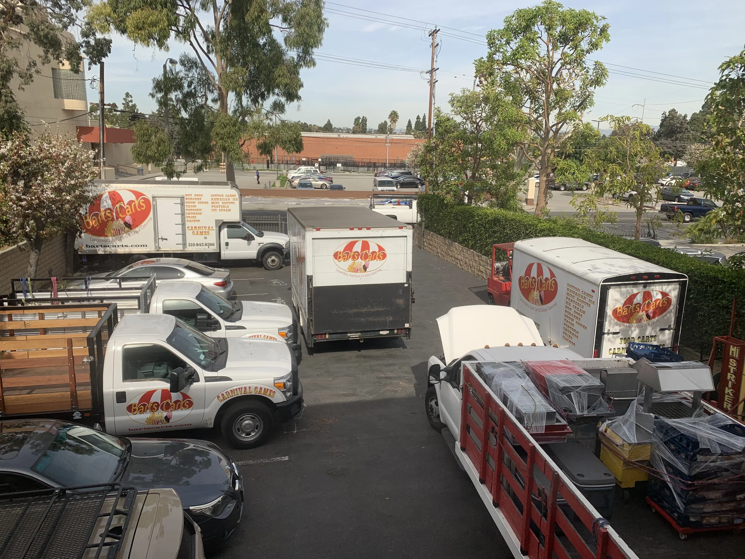 Parking lot with trucks and cars, trees, and a building in the background under a partly cloudy sky.