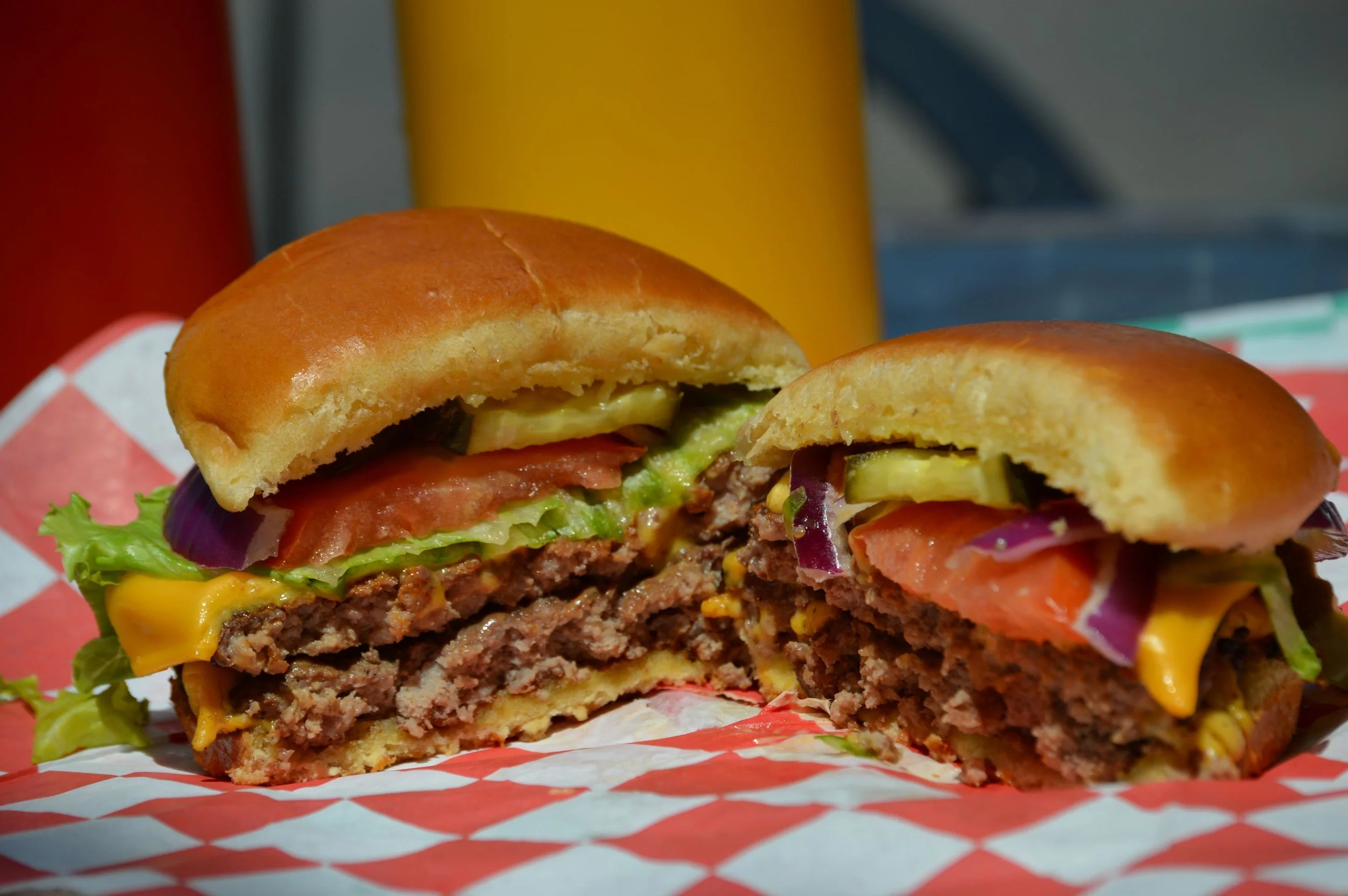 Close-up of a half-eaten cheeseburger with lettuce, tomato, pickles, onion, melted cheese, hamburger patty, and a bun, on red checkered paper with yellow, red, and gray cans in the background.