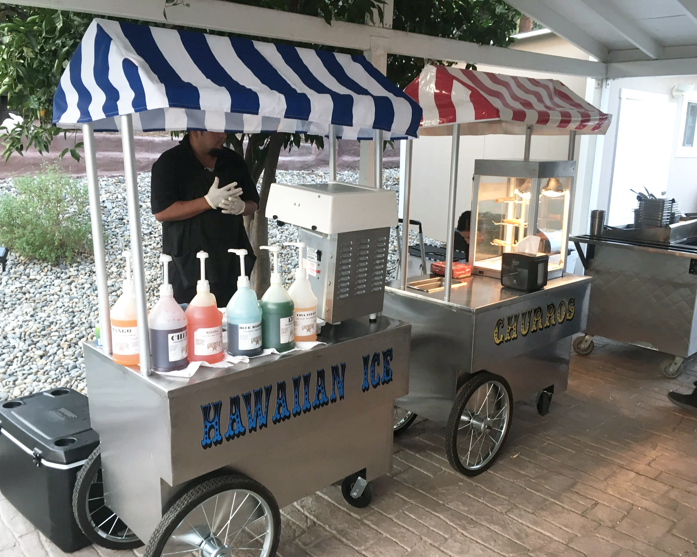 A snow cone stand cart with a striped canopy, bottles of flavored syrup on a table, and a worker preparing snow cones. The cart has text indicating it serves Hawaiian Ice and Churros.